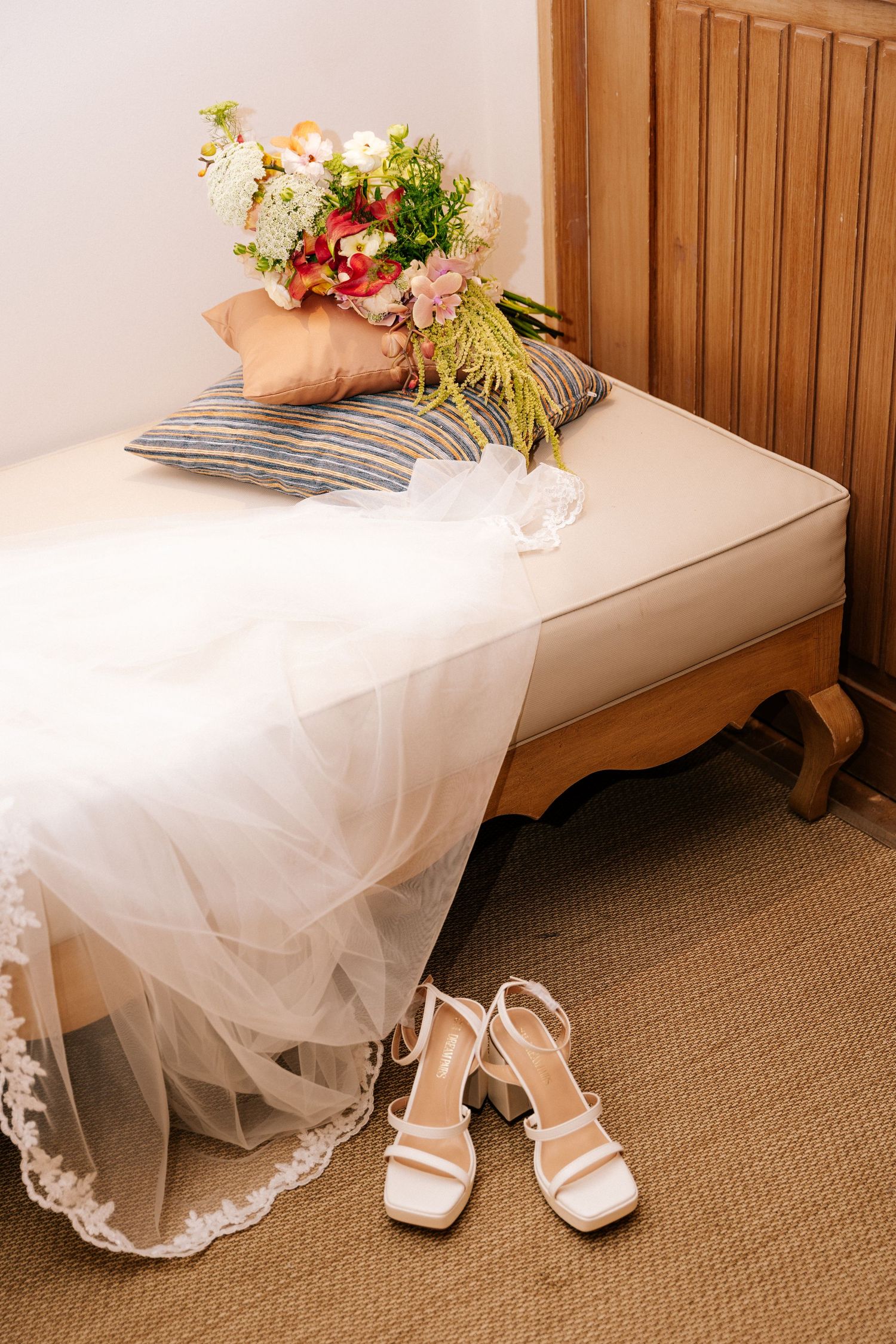 Bridal wedding setup with white heels, lace gown, floral bouquet, and pillows on a wooden bench.