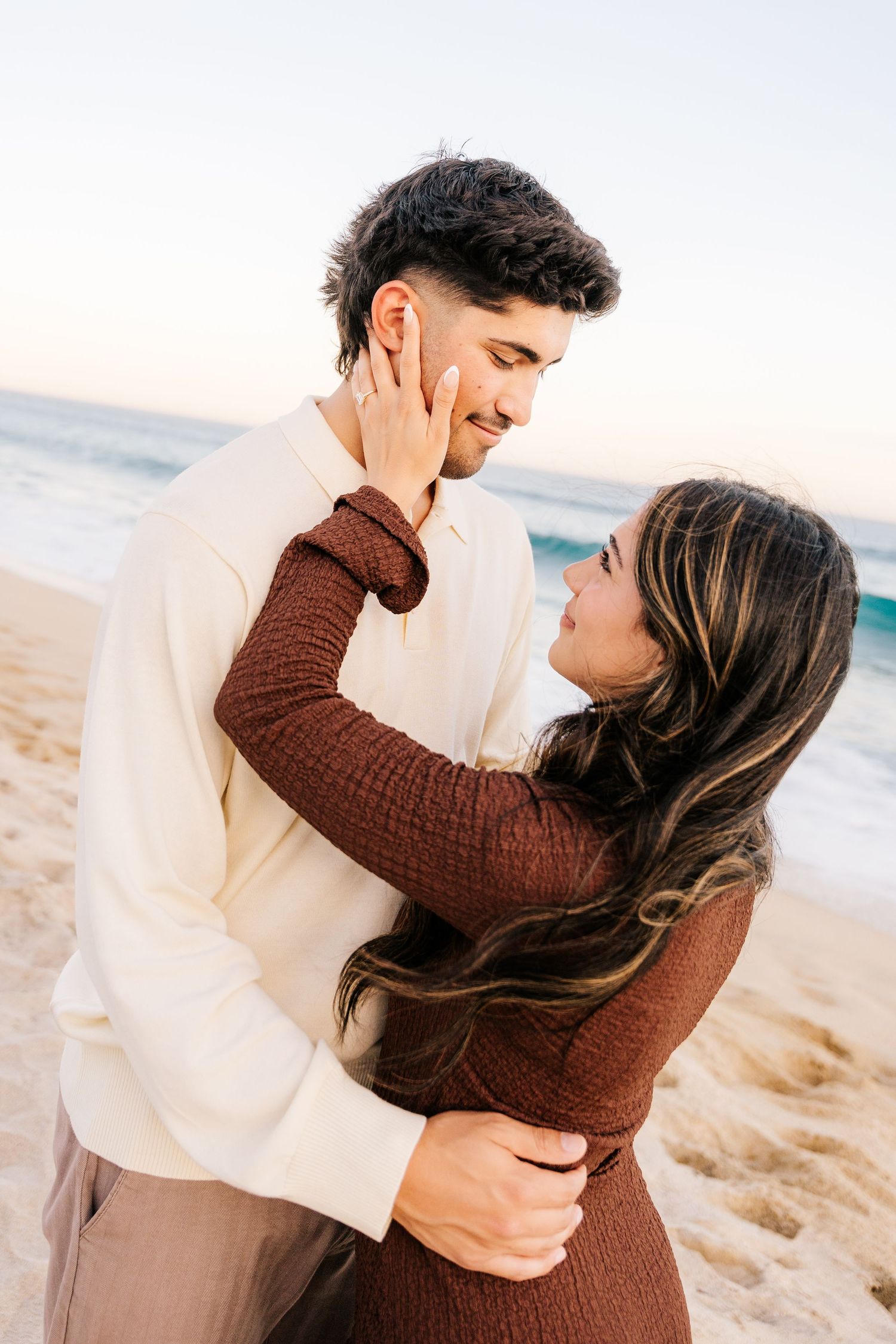 Romantic couple embracing on a sandy beach at sunset, woman in brown sweater touching man's face tenderly.