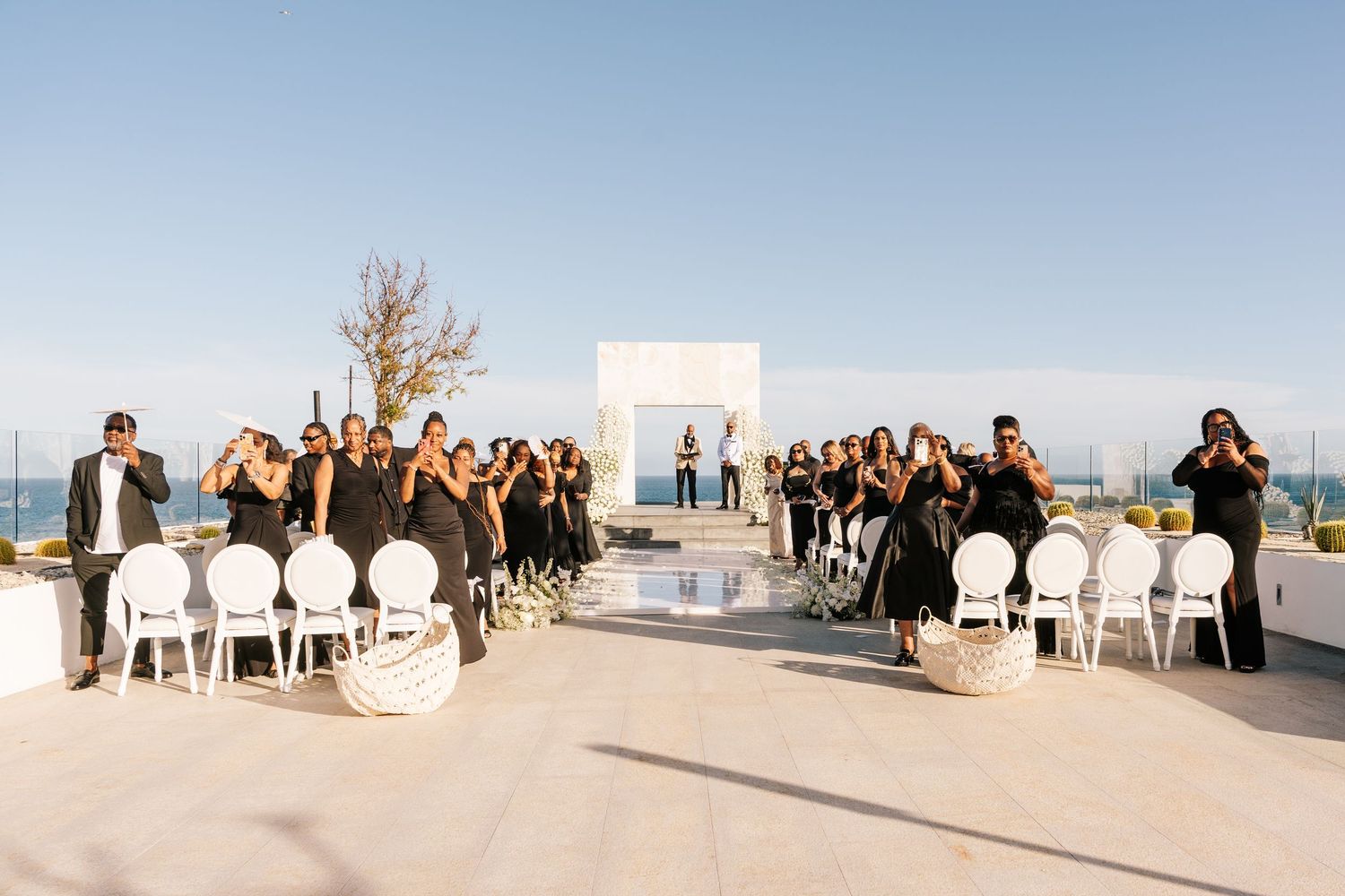 Outdoor rooftop wedding ceremony with guests in black attire, white chairs, and a minimalist altar under a clear blue sky.