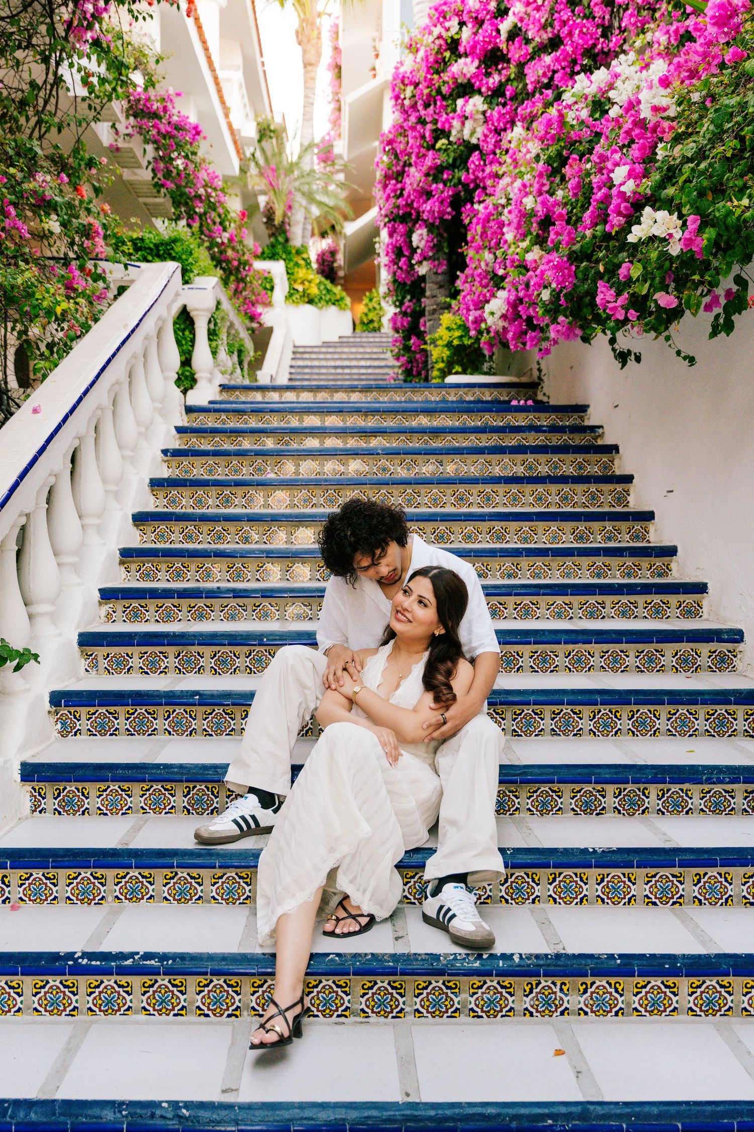 Couple sitting on decorative tiled stairs surrounded by vibrant pink bougainvillea flowers in a romantic outdoor setting.