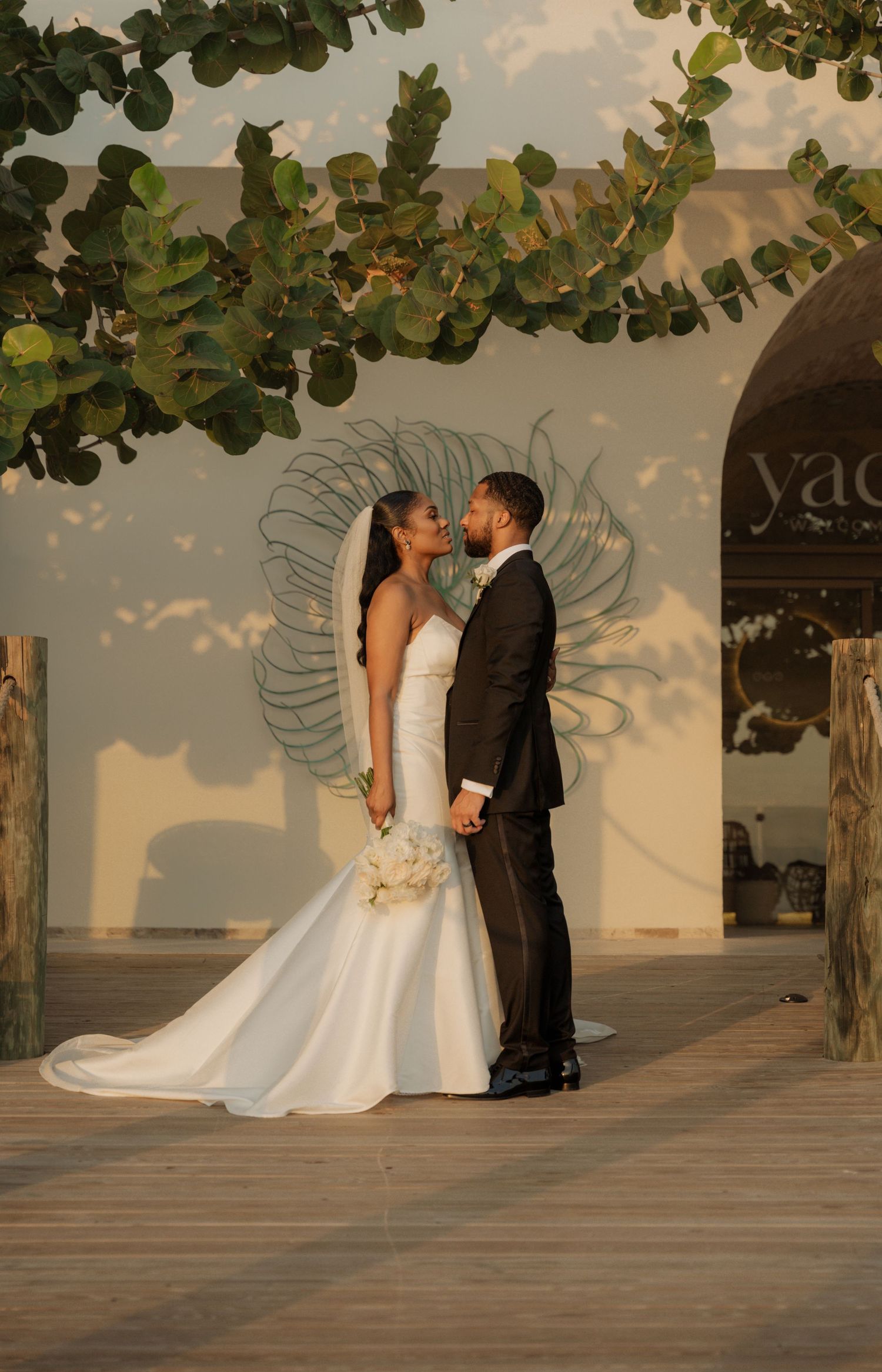 A romantic wedding couple embraces at sunset under green foliage and a decorative arch.