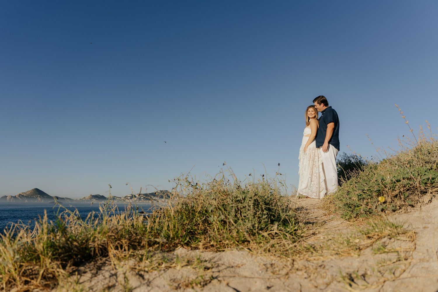 Couple embracing on sandy coastal dune with blue ocean and clear sky backdrop during golden hour.
