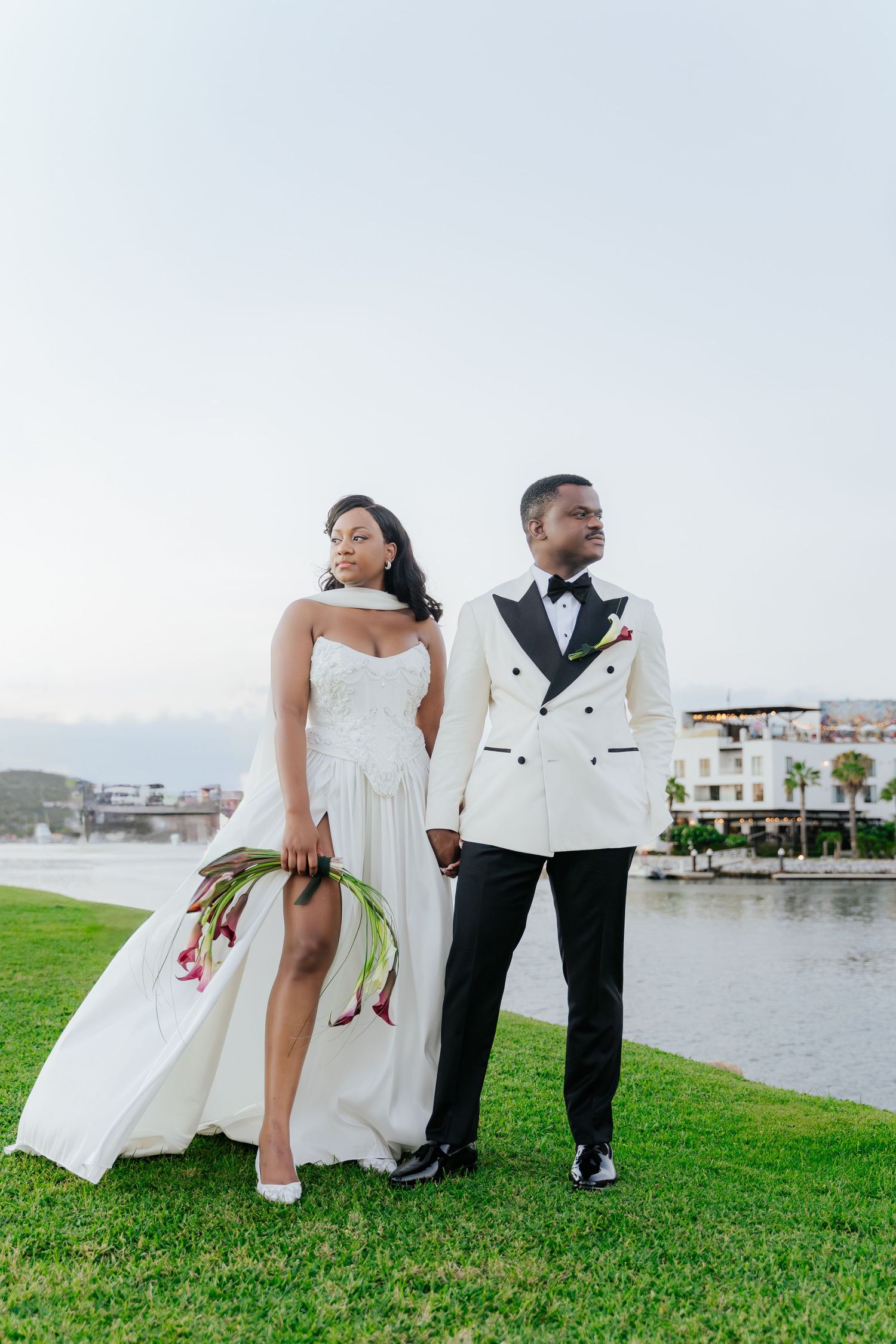 A couple in wedding attire sits together on green grass by the waterfront during their special day.