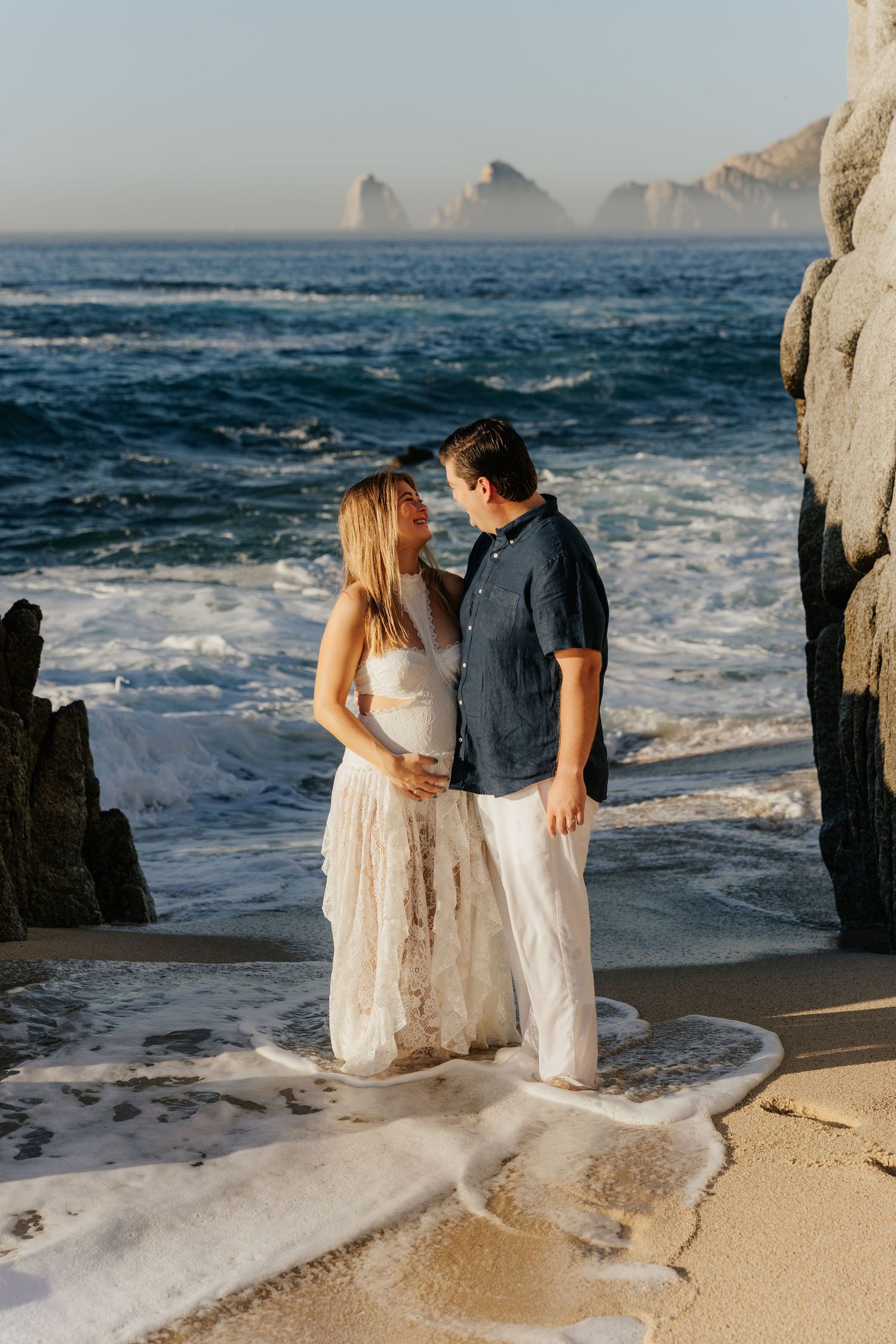 Couple sharing a romantic moment on a sandy beach at sunset with ocean waves and rocky cliffs in Cabo.