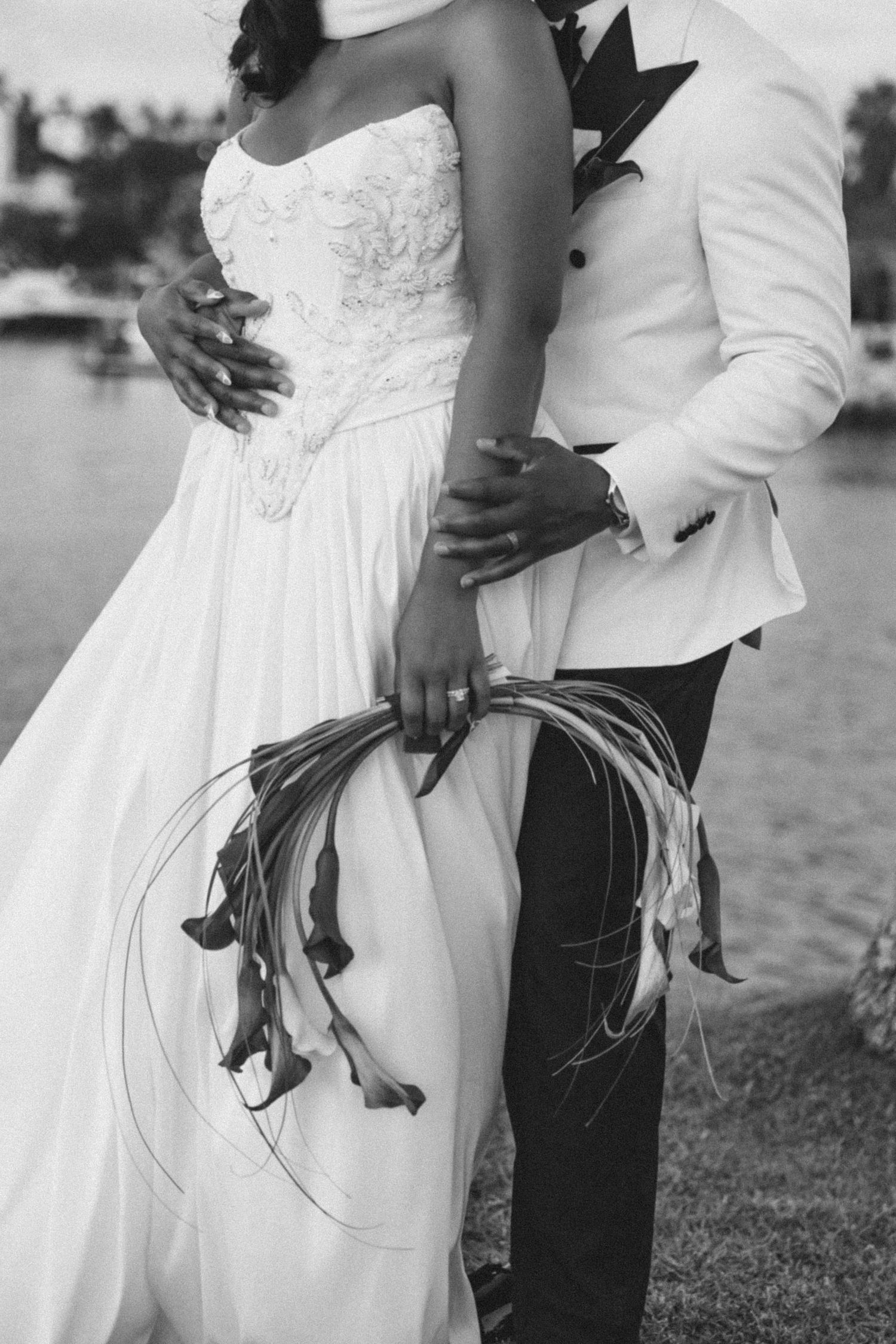 Black and white wedding photo showing couple embracing outdoors, with focus on their torsos in elegant white attire.