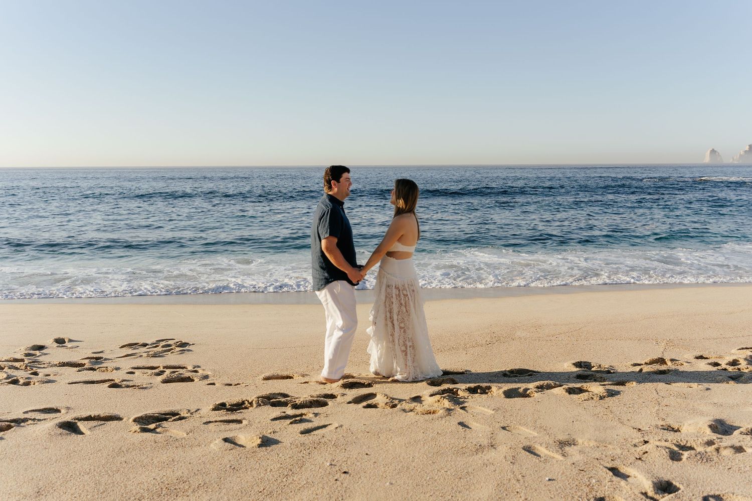 Couple holding hands on a sandy beach with ocean waves in the background during golden hour.