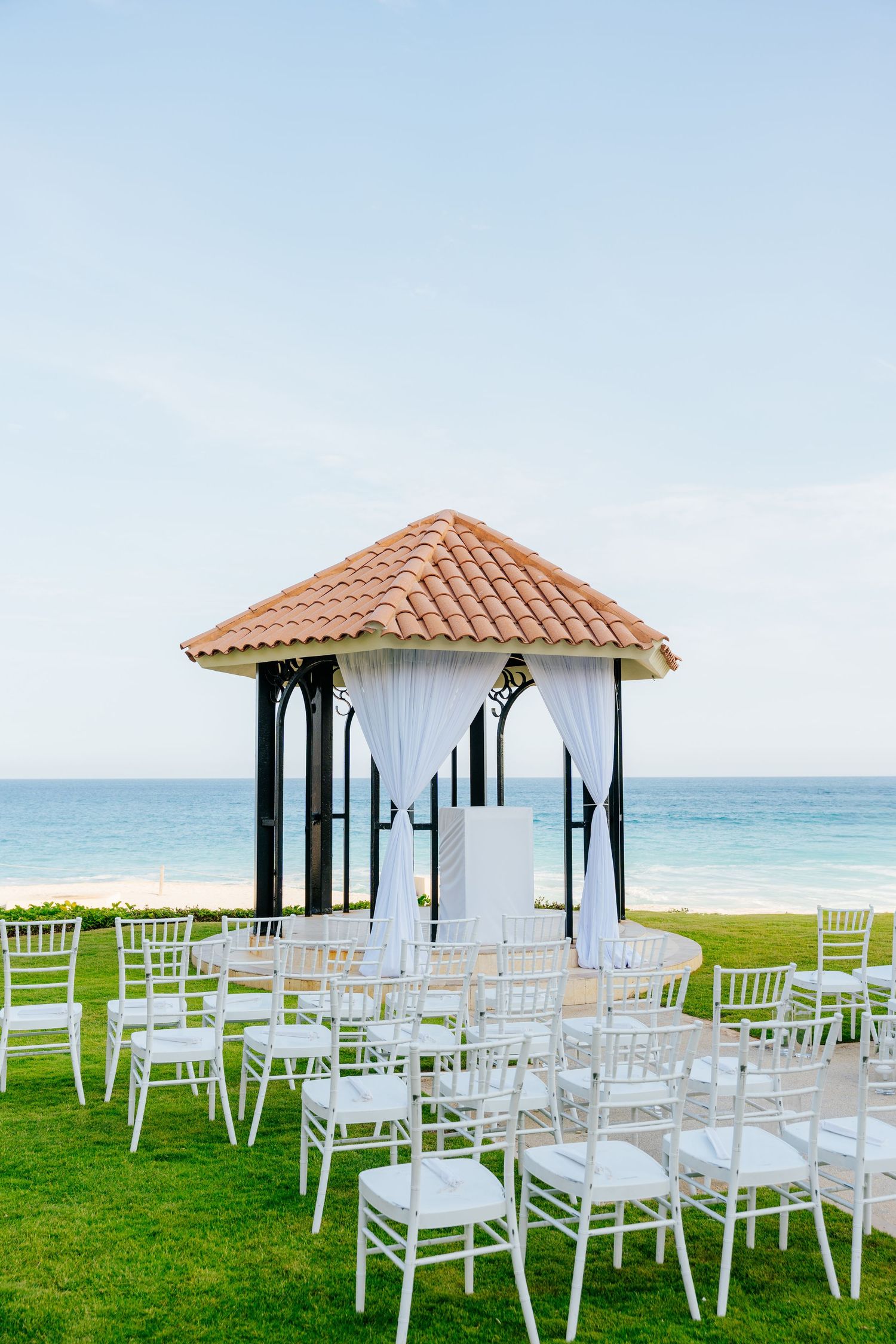 Oceanfront wedding gazebo with terracotta roof and white drapes, surrounded by white chairs on a green lawn overlooking the beach.
