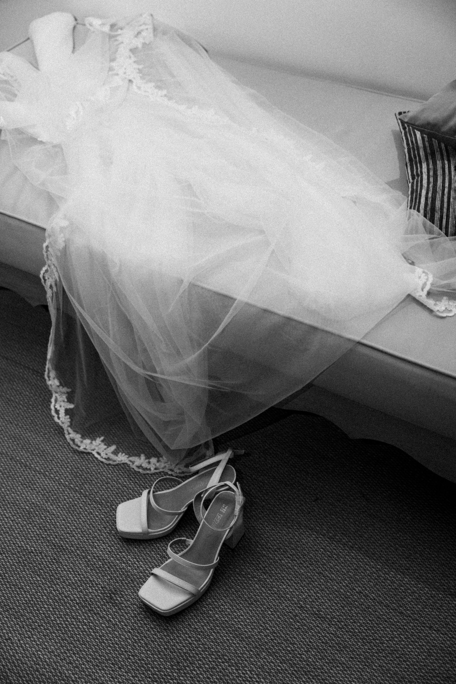 Black and white photo of a lace-trimmed wedding dress and veil draped over a sofa, with white block-heel sandals on the floor below.