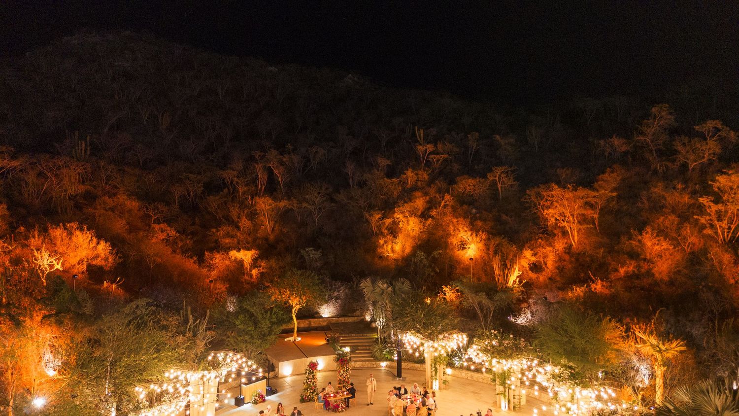 Panoramic aerial view of an illuminated outdoor festival with glowing tents and trees at night.