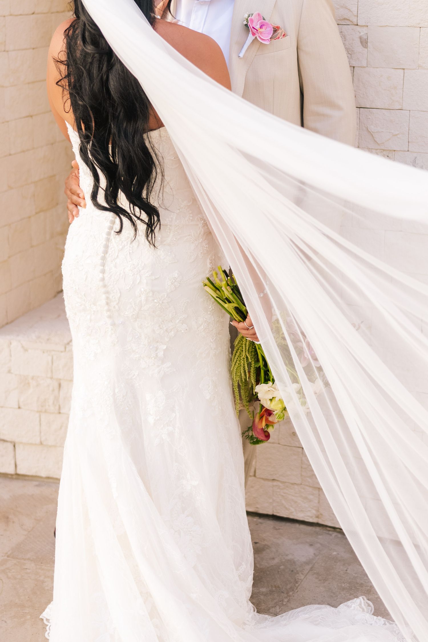Bride in lace mermaid gown with flowing veil and bouquet embracing groom in beige suit at outdoor wedding.