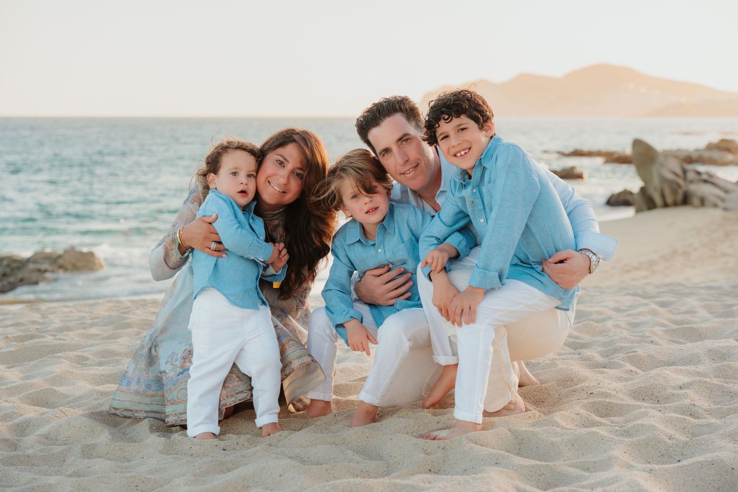 Family wearing matching blue and white outfits poses together on a sandy beach at sunset.