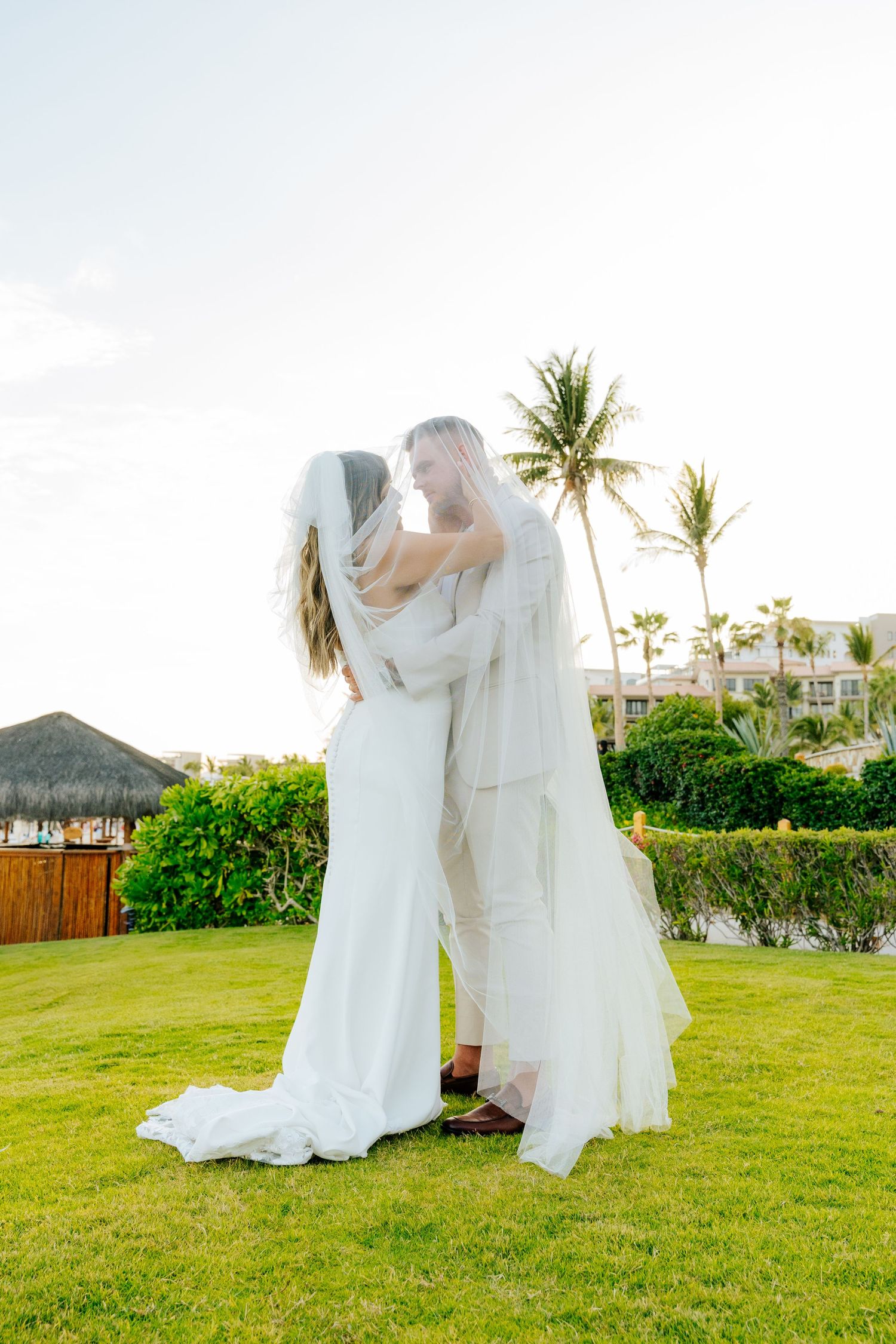 Bride and groom embrace under a sheer veil on a lush green lawn at a tropical beach wedding with palm trees and a thatched hut.