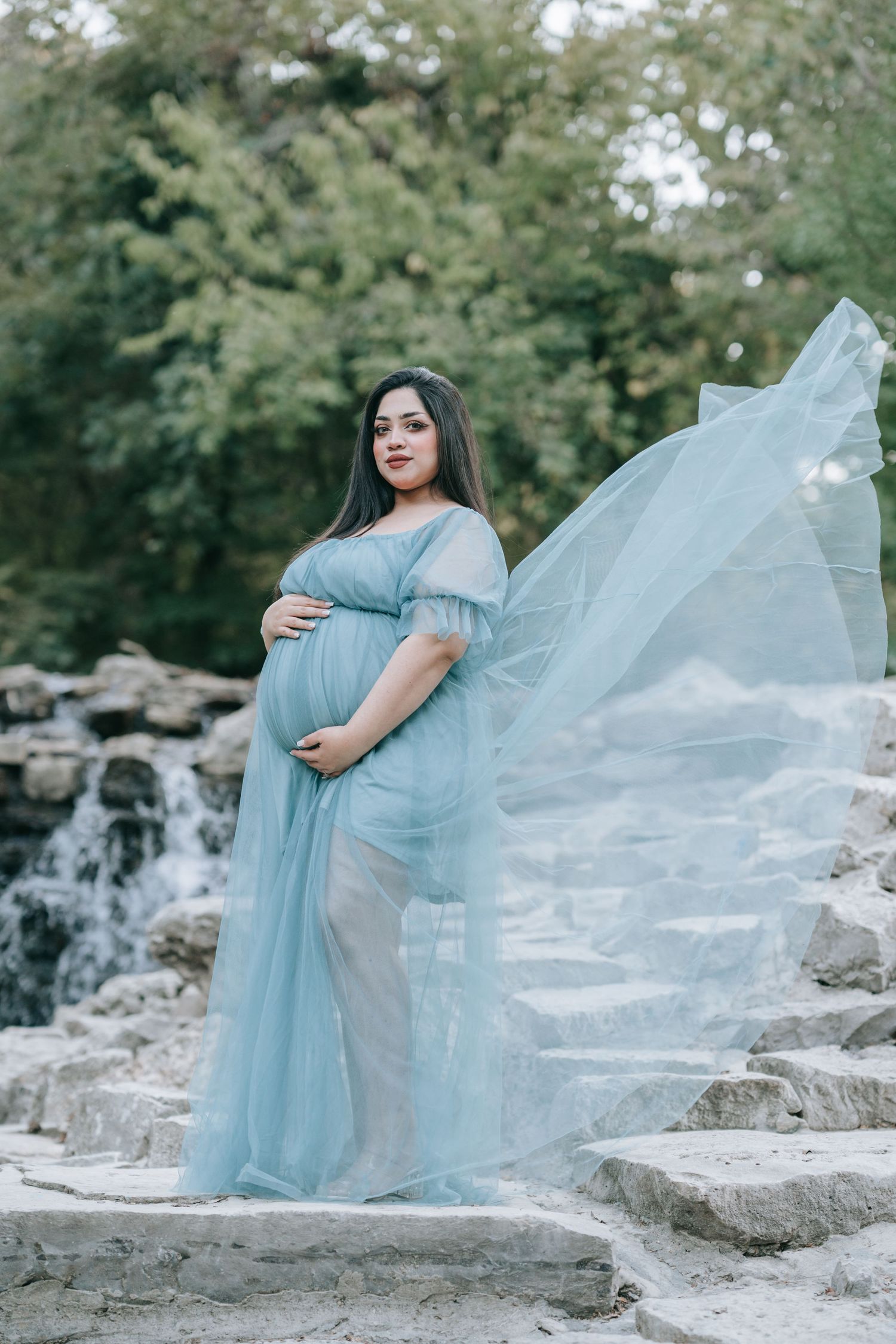 A maternity photo session featuring a flowing light blue dress against a cascading waterfall backdrop.