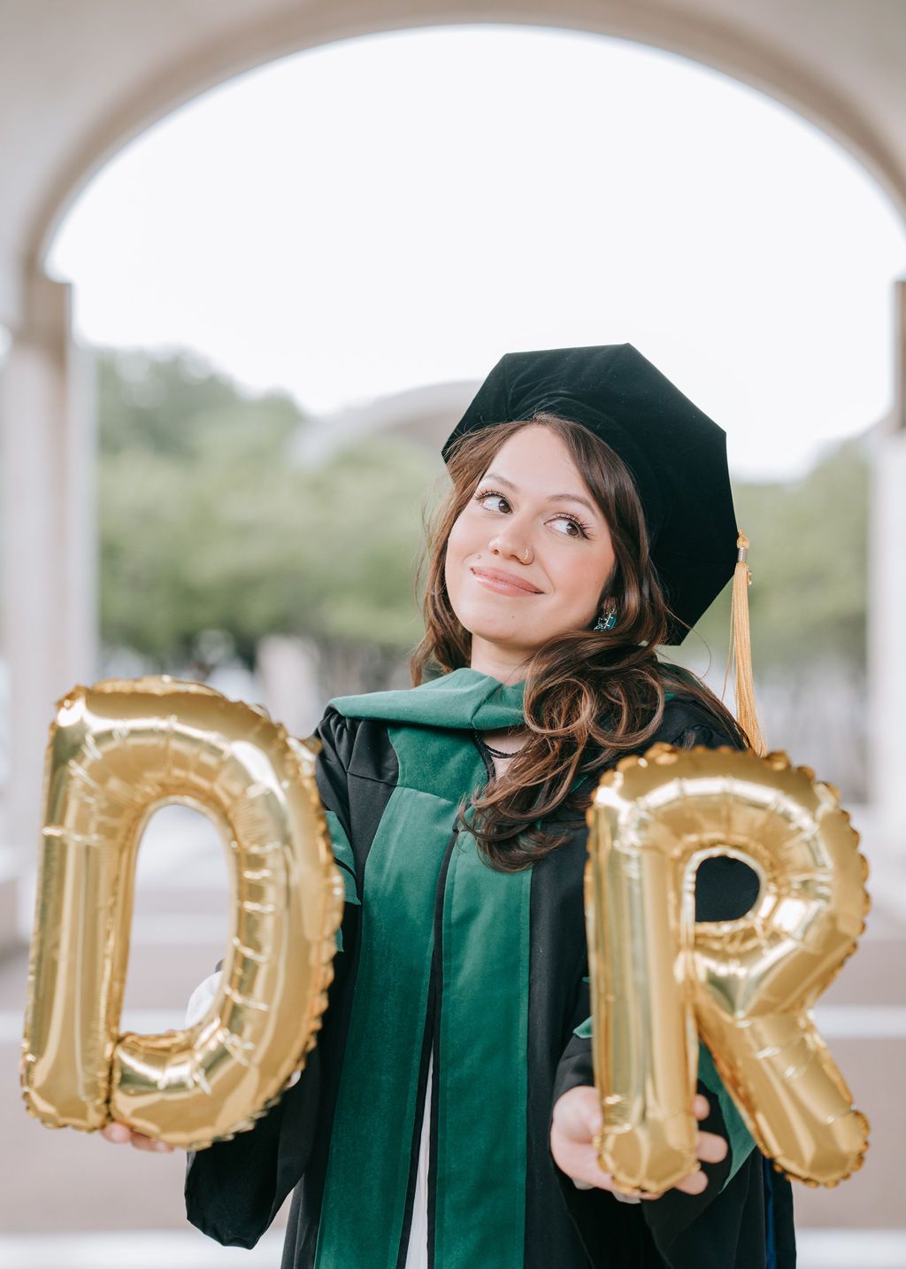 Graduate in green doctoral regalia poses with gold letter balloons under archway.