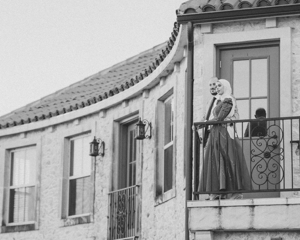 Person in vintage dress standing on ornate balcony of curved Mediterranean-style building.