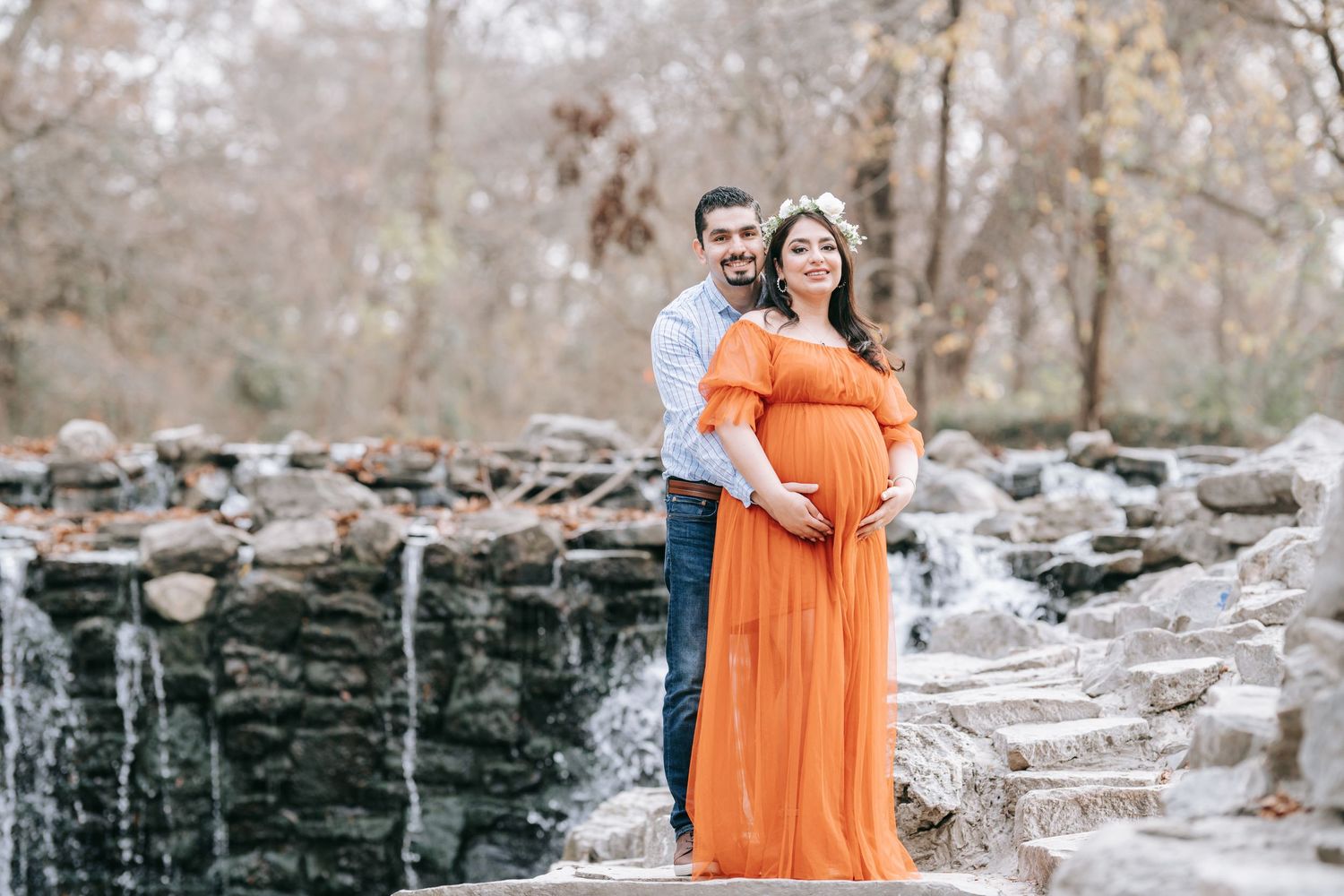 A maternity photoshoot by a waterfall with a woman wearing a vibrant orange dress.