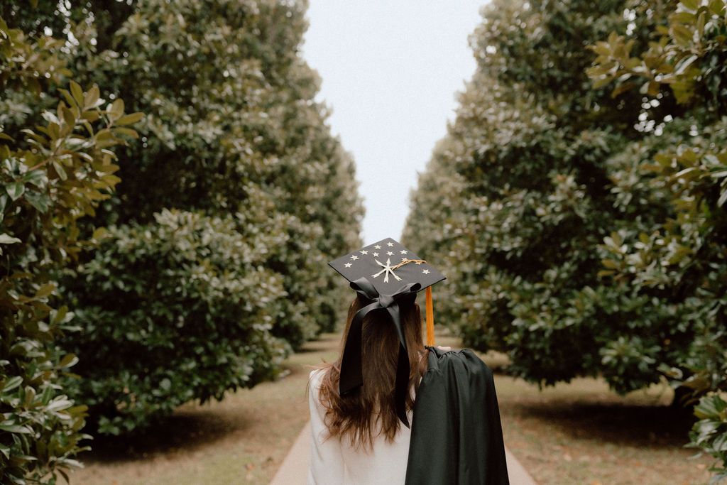 A graduate in cap and gown walking down a tree-lined path.