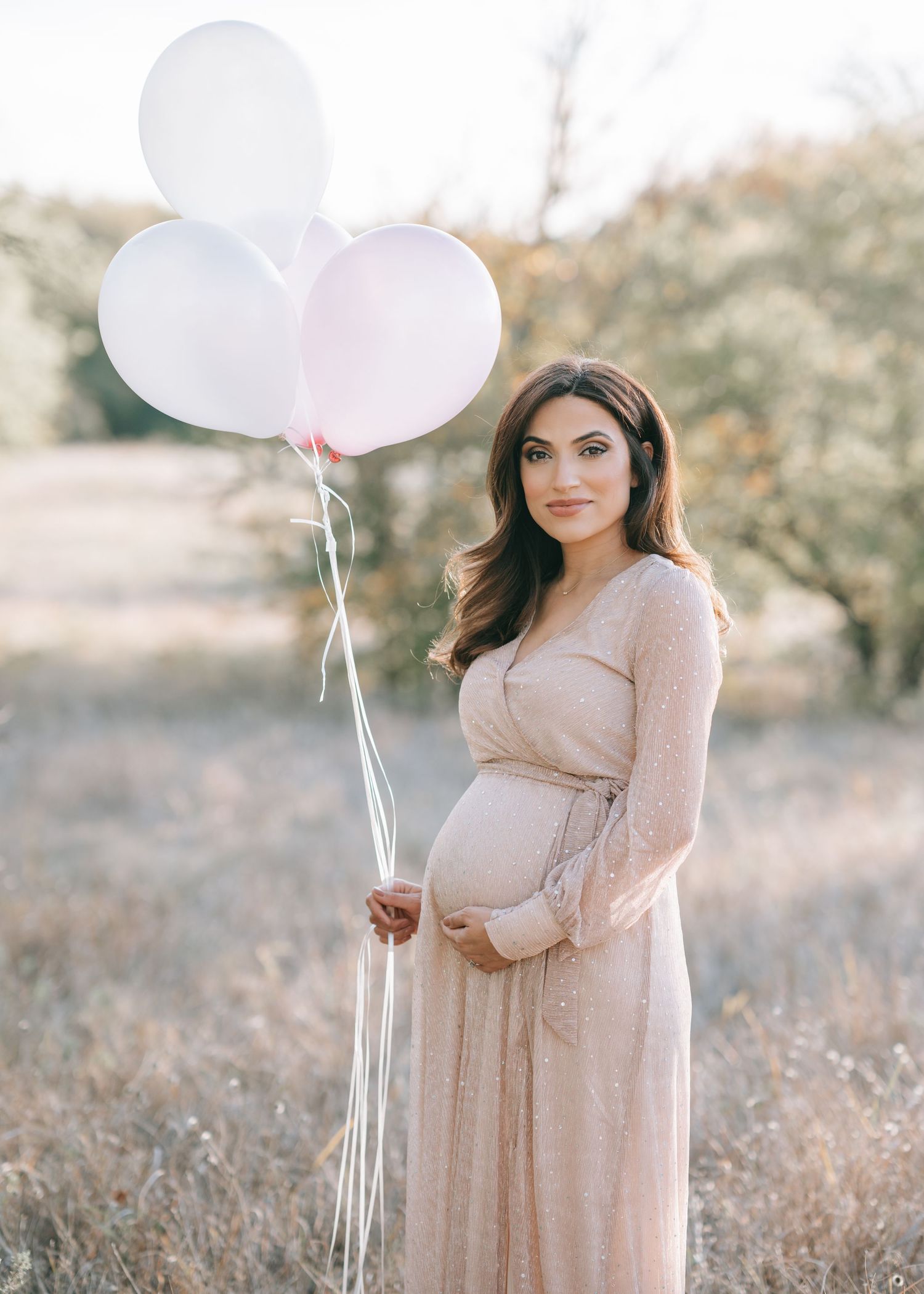 A pregnant woman in a blush dress holds white and pink balloons in a dreamy outdoor setting.