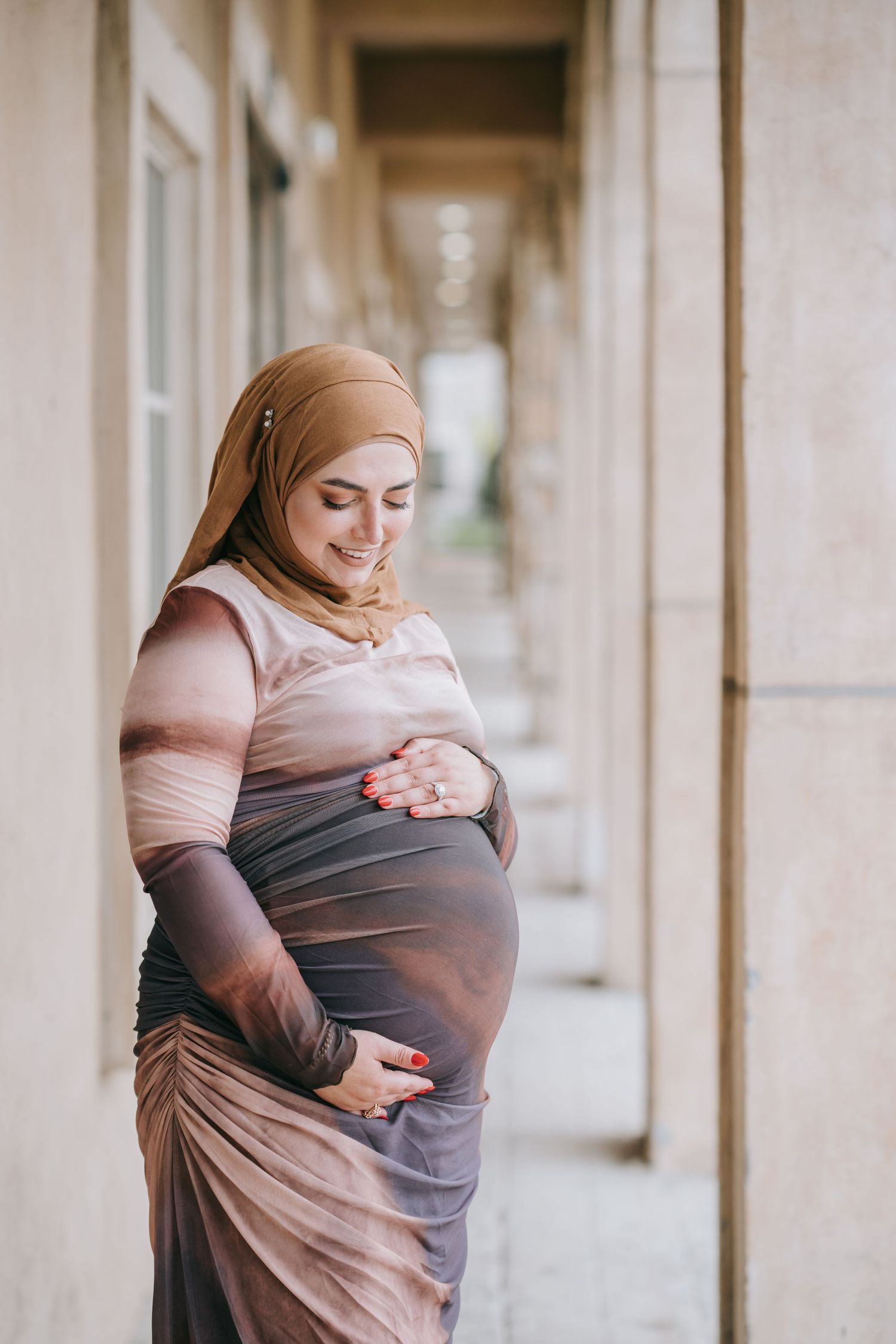 Maternity portrait of woman in hijab and brown dress posing in columned hallway.