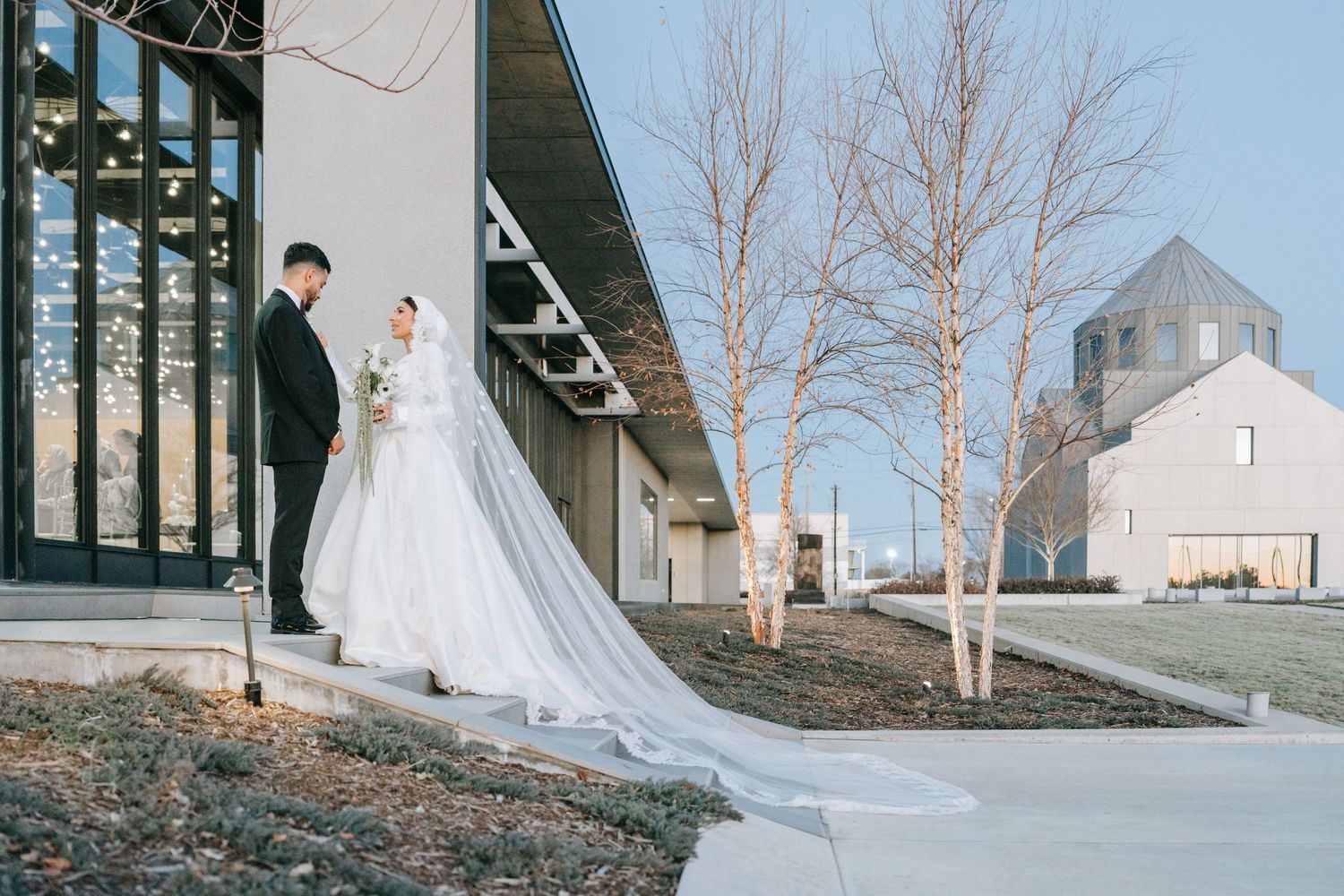 A couple in wedding attire stands outside a modern building with a white church visible in the background.