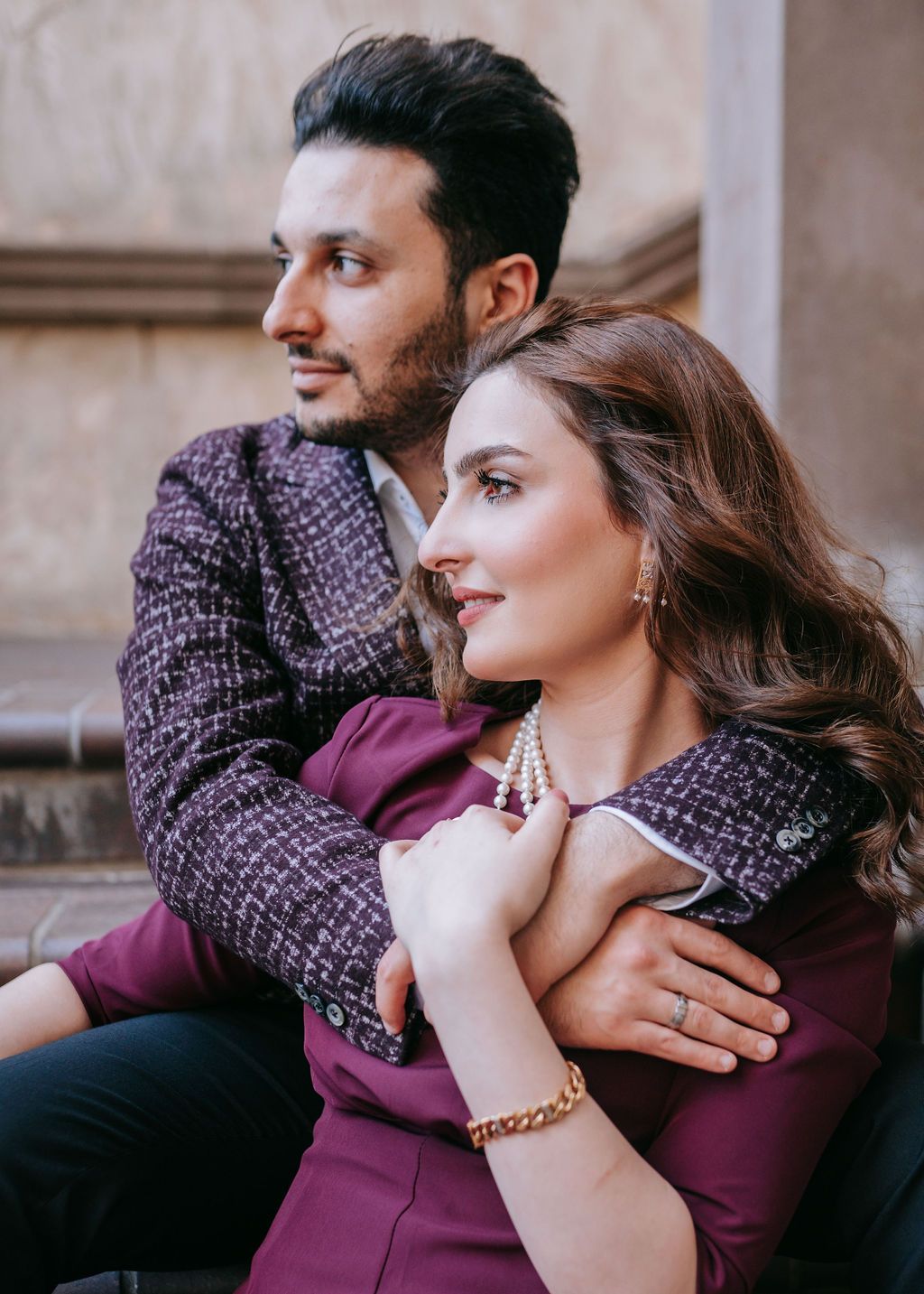 Couple in coordinating purple outfits pose elegantly against stone wall.