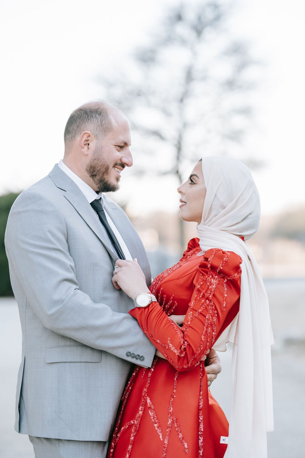Couple in formal attire sharing romantic moment outdoors, with red dress and light grey suit.