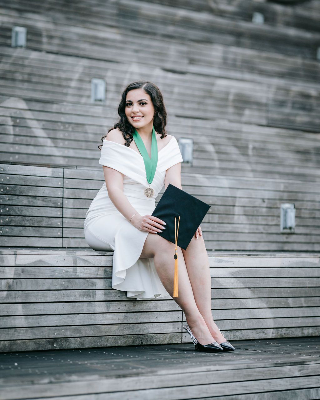 Graduate in white dress and green stole sits on wooden stadium steps holding diploma cover.