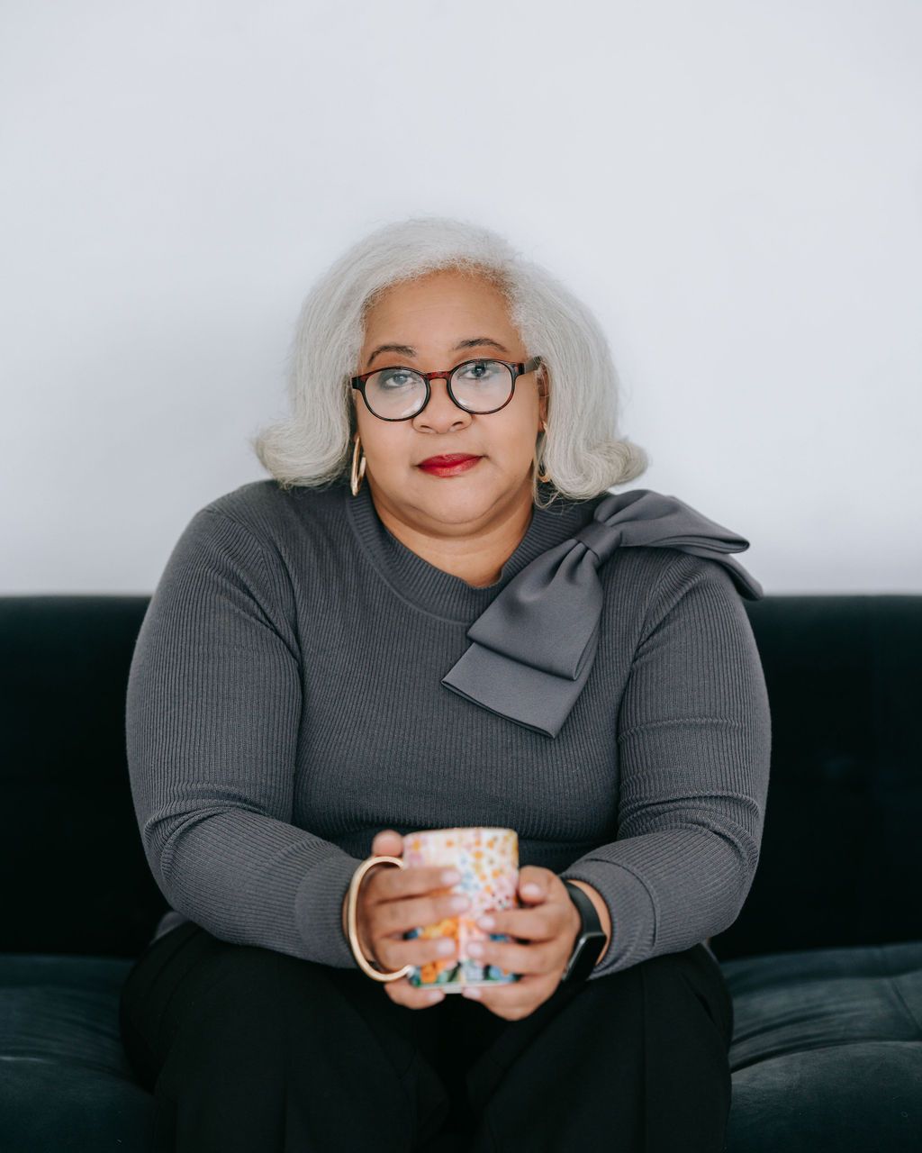 Woman in grey sweater with silver hair and glasses holds coffee mug while sitting on couch.