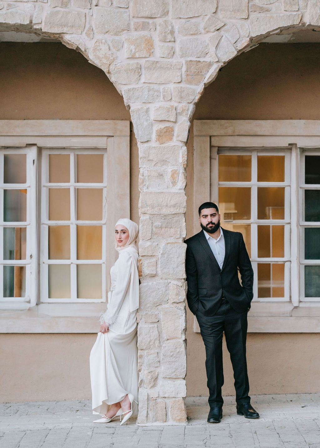 A formal couple poses separately against stone archways in traditional wedding attire.