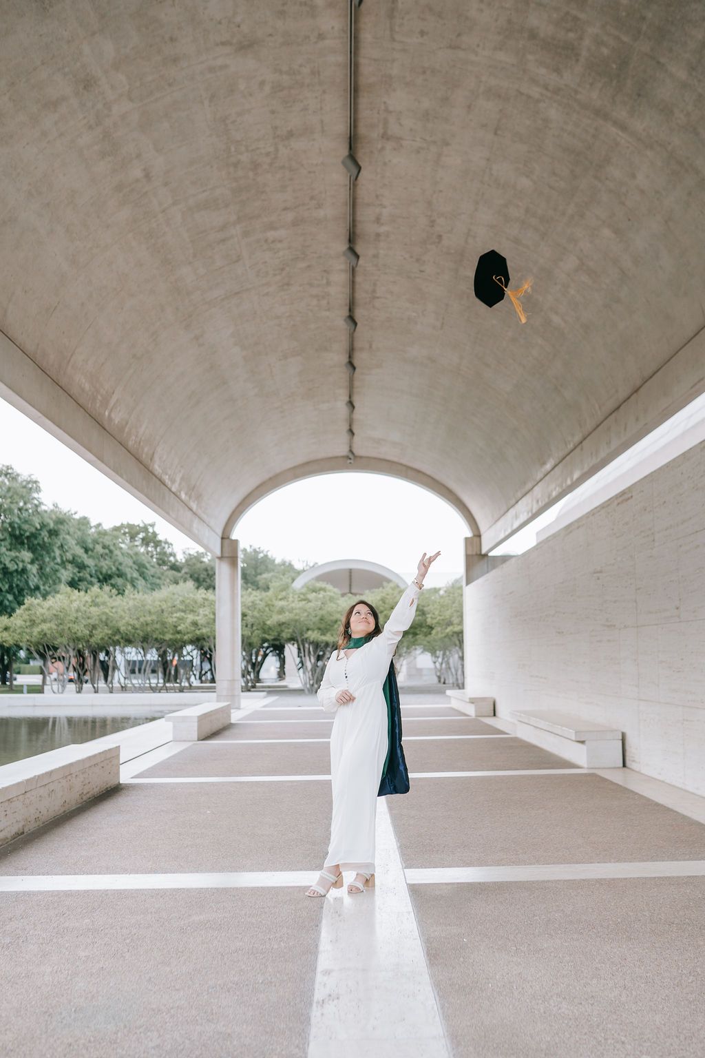 Graduate in flowing white dress celebrates under concrete archway near reflecting pool.