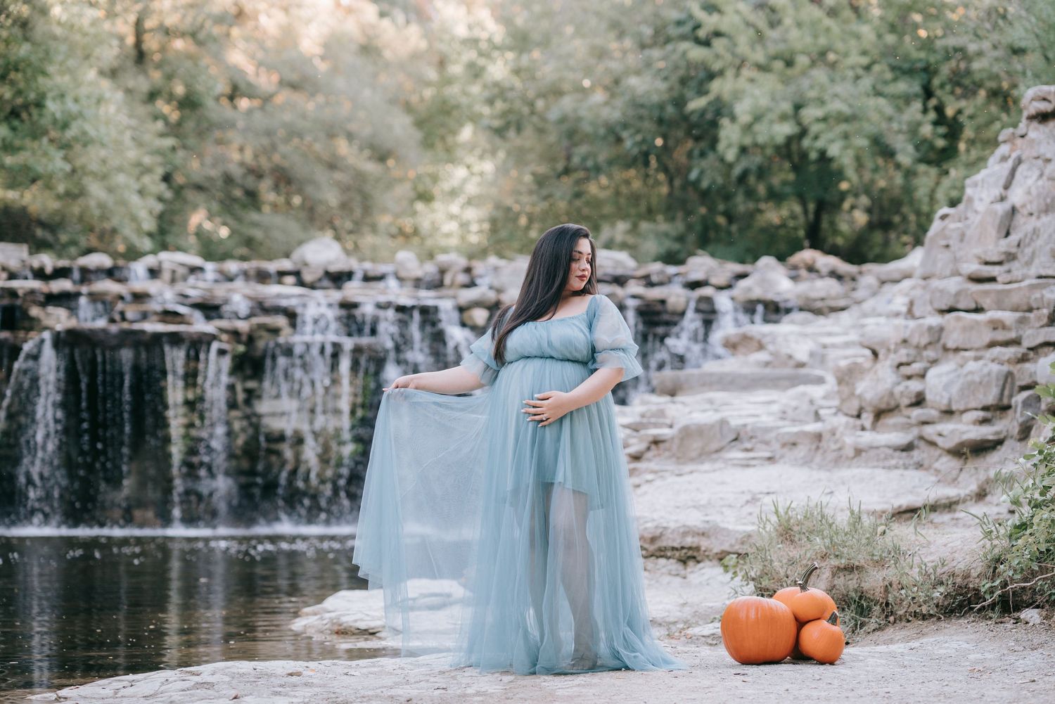 A maternity photoshoot by a waterfall with a light blue dress and pumpkin decorations.