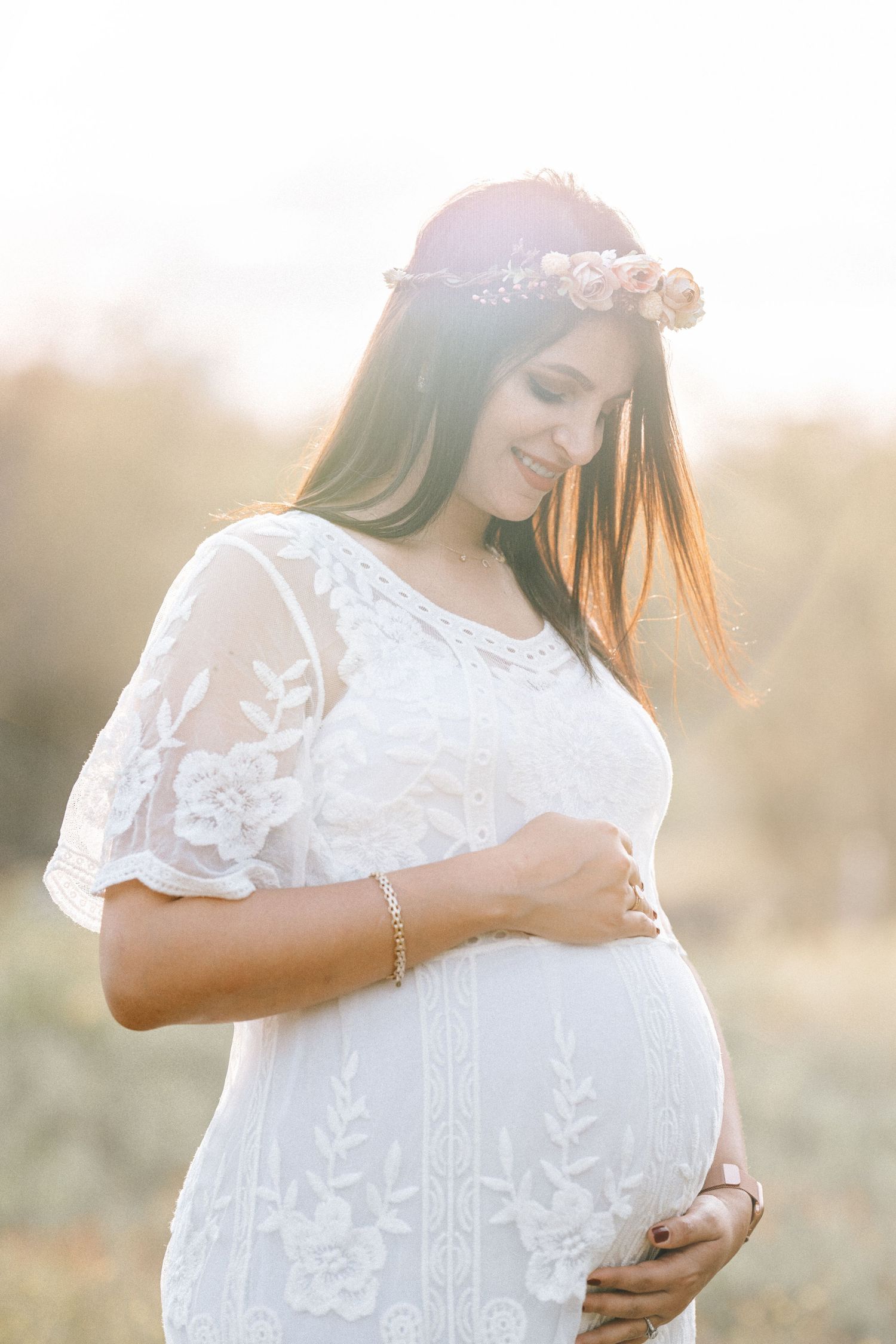 A pregnant woman in a white lace dress and flower crown smiles while touching her belly in golden sunlight.