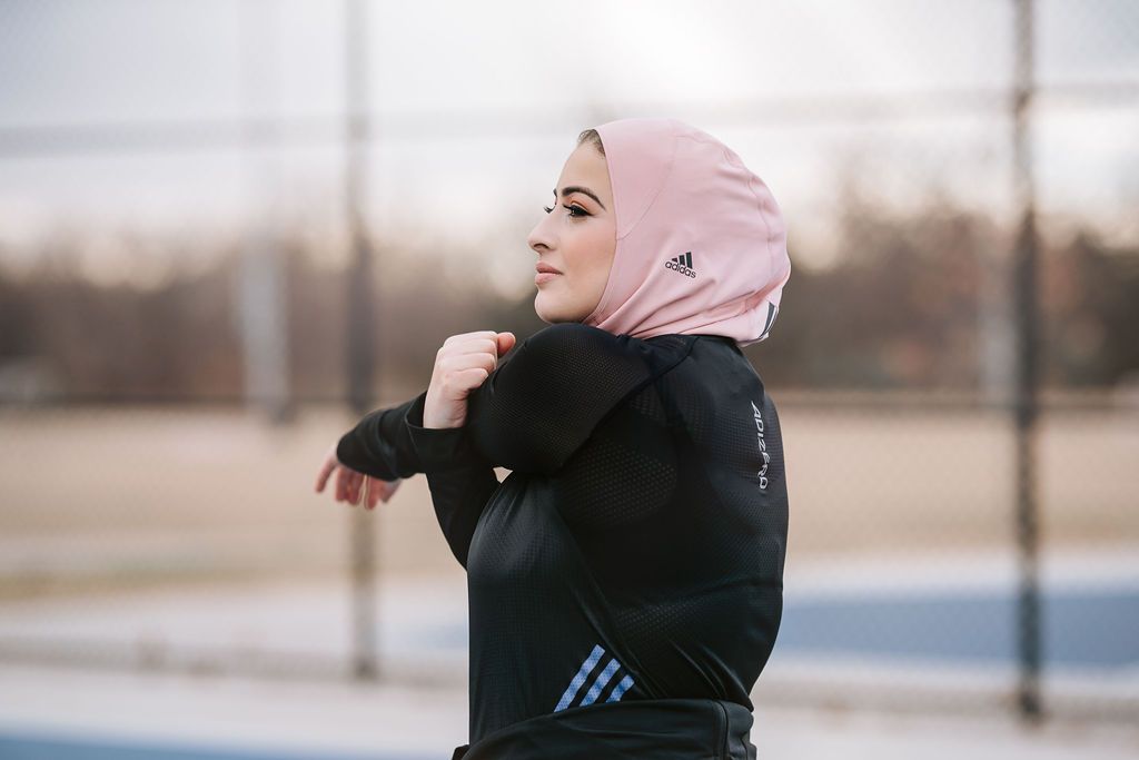 Person in black athletic wear and pink hijab stretching outdoors.