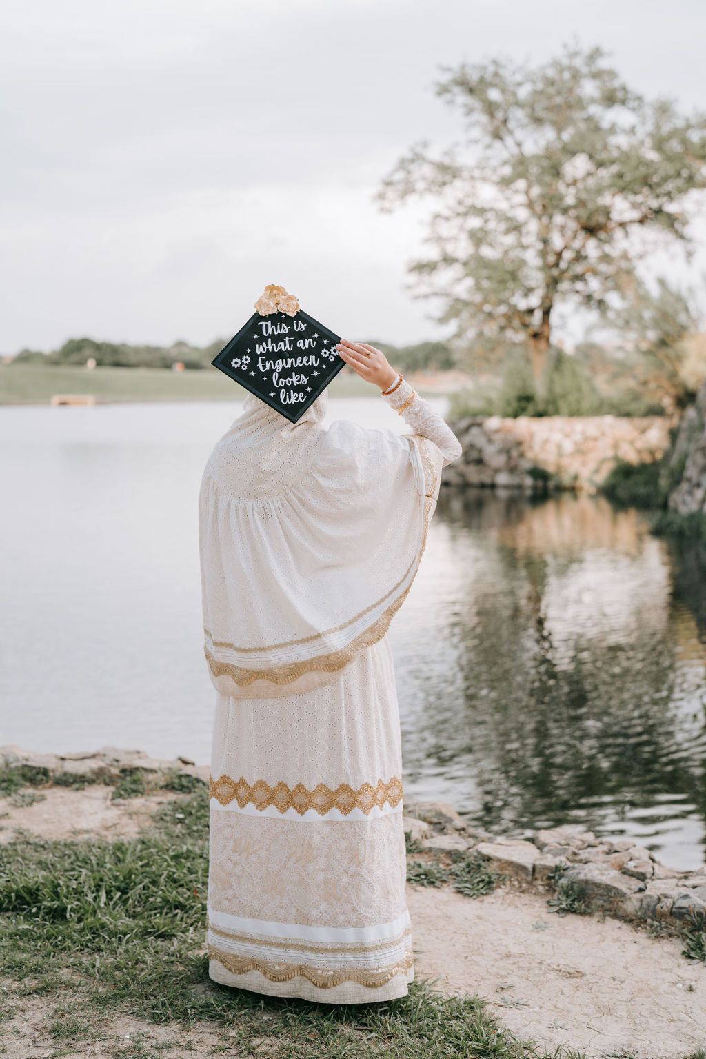 A graduate in white dress holds up decorated cap by a reflective pond.