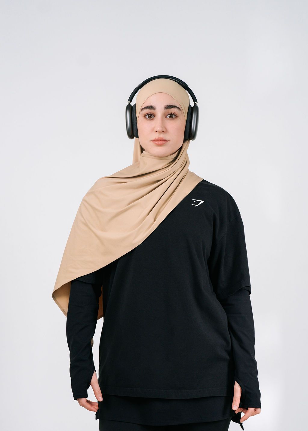 Person wearing a black sweater, tan hijab, and over-ear headphones against a light background.