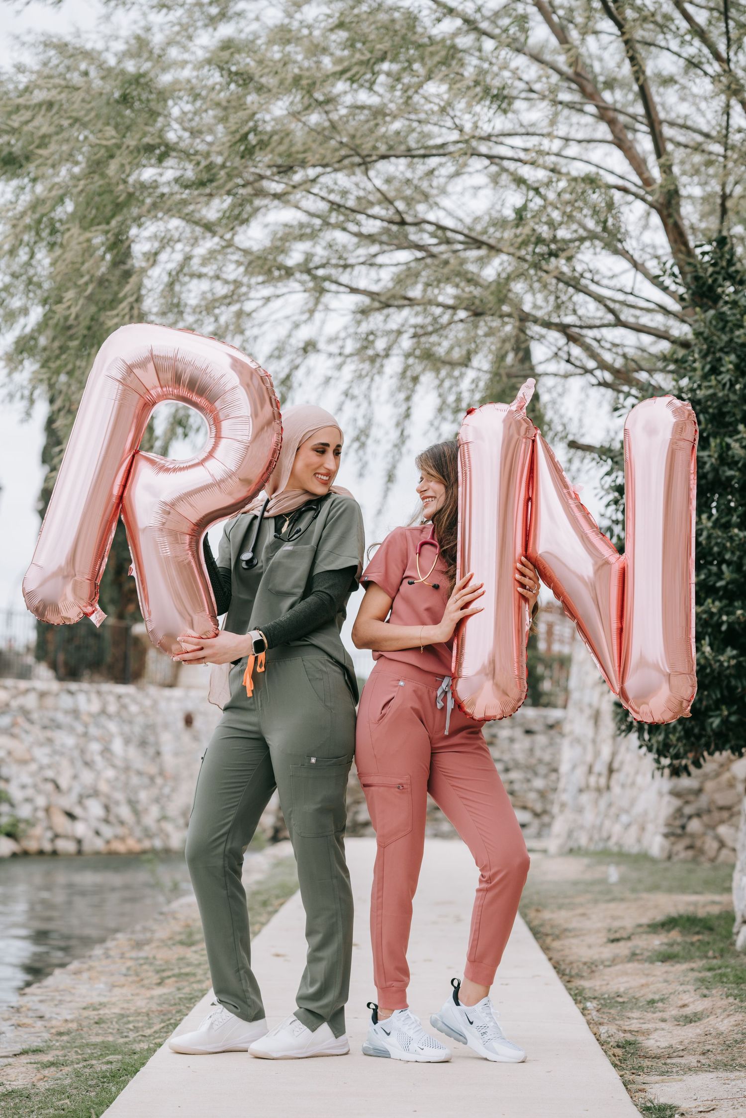 Two people posing with large rose gold letter balloons spelling 'RN' by a waterfront path.
