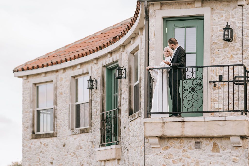 A couple standing on a Mediterranean-style balcony with stone facade and green shutters.