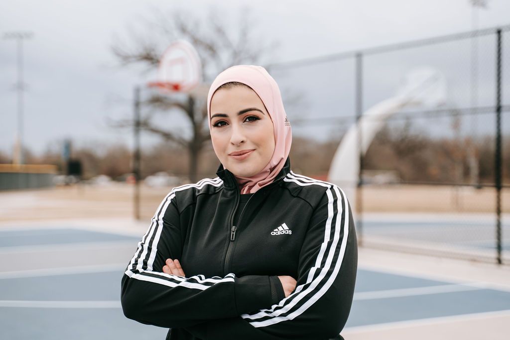 Athletic figure in Adidas jacket and hijab poses confidently on outdoor basketball court.
