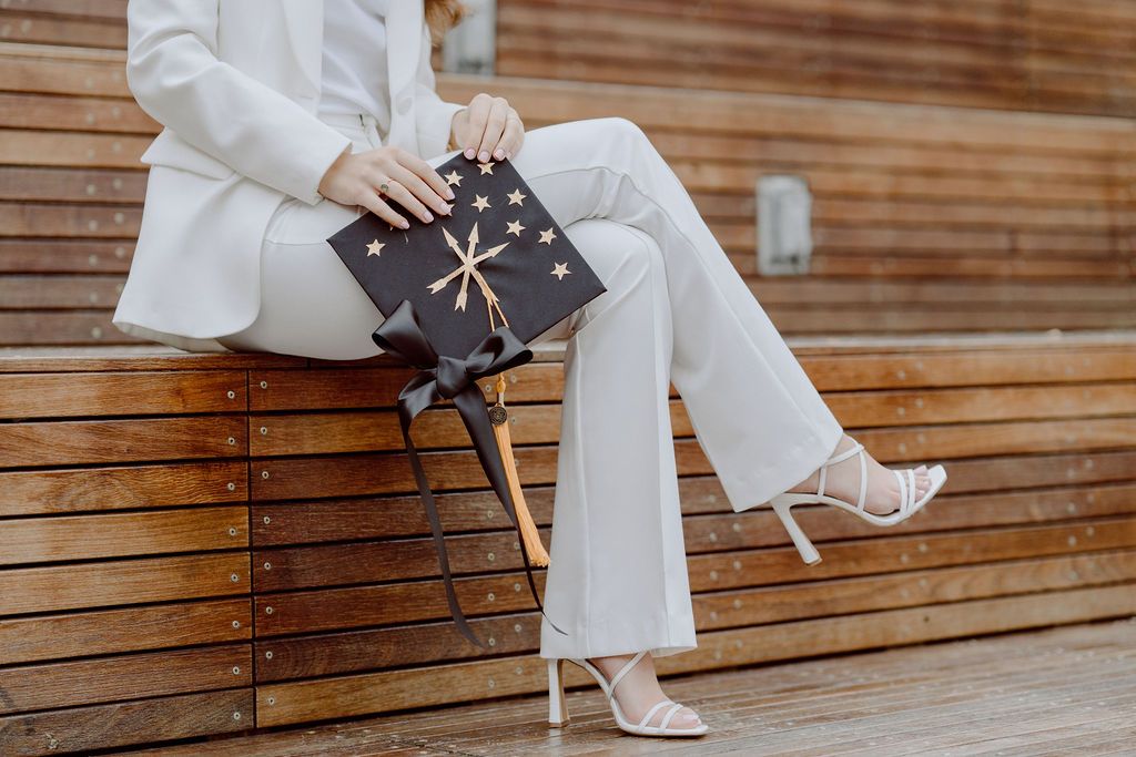 Graduate in white outfit sits on wooden bench displaying decorated graduation cap.