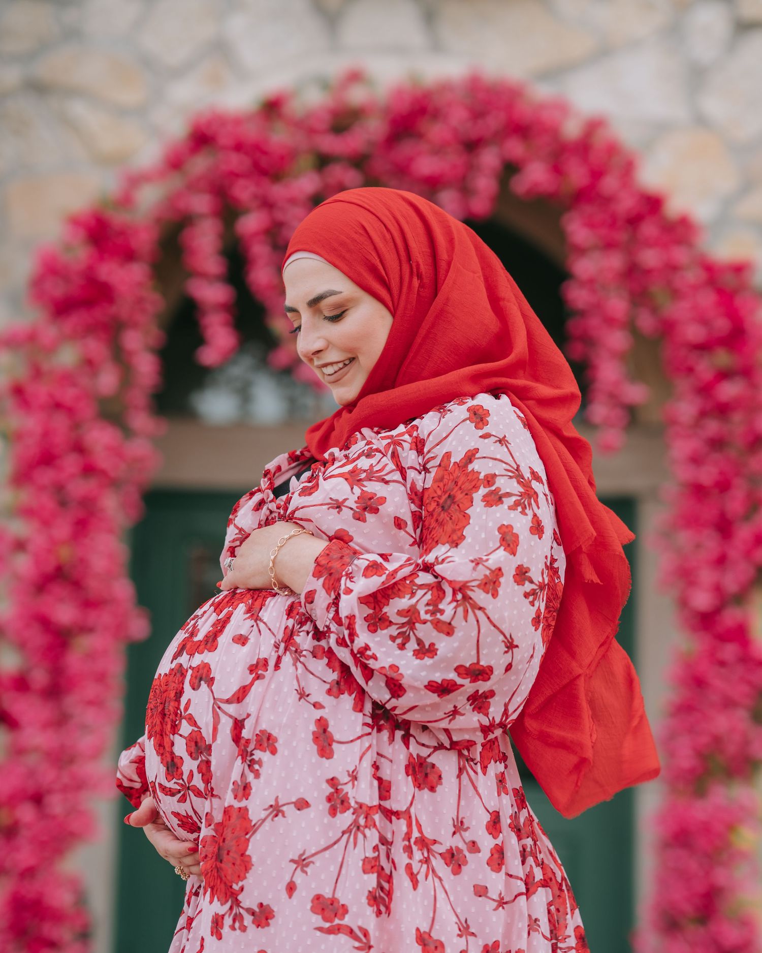 Expectant mother in red hijab and floral dress poses under pink floral arch during maternity shoot.