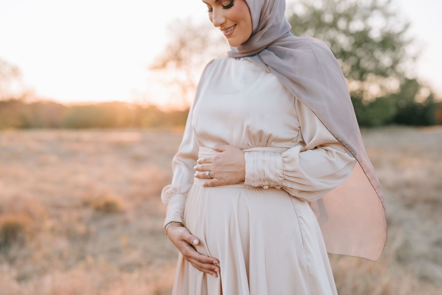 A pregnant woman in a flowing white dress poses at sunset in nature.