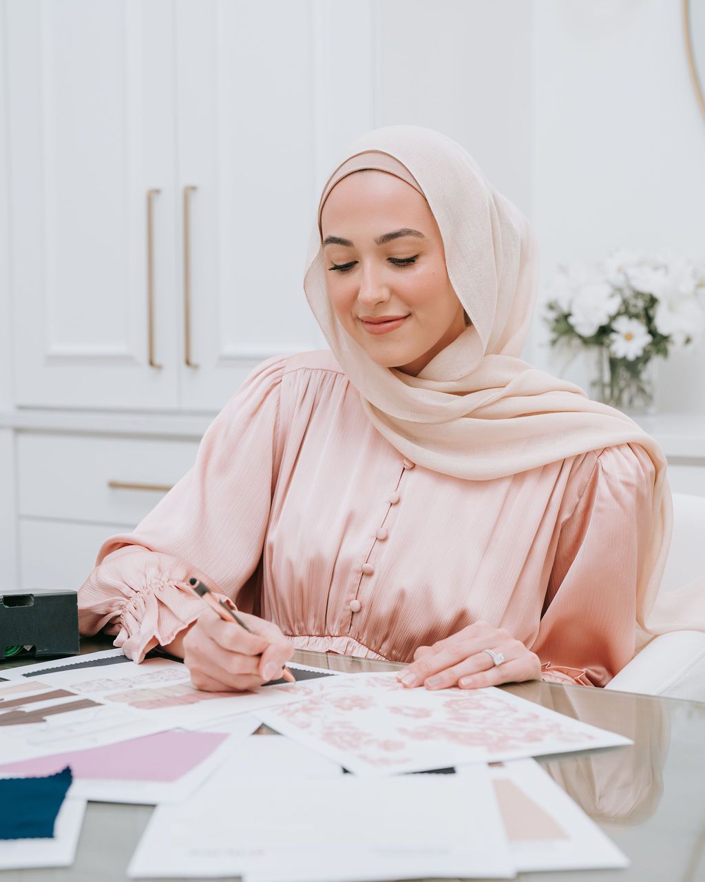 Person in pink hijab writing at desk with office supplies and papers.