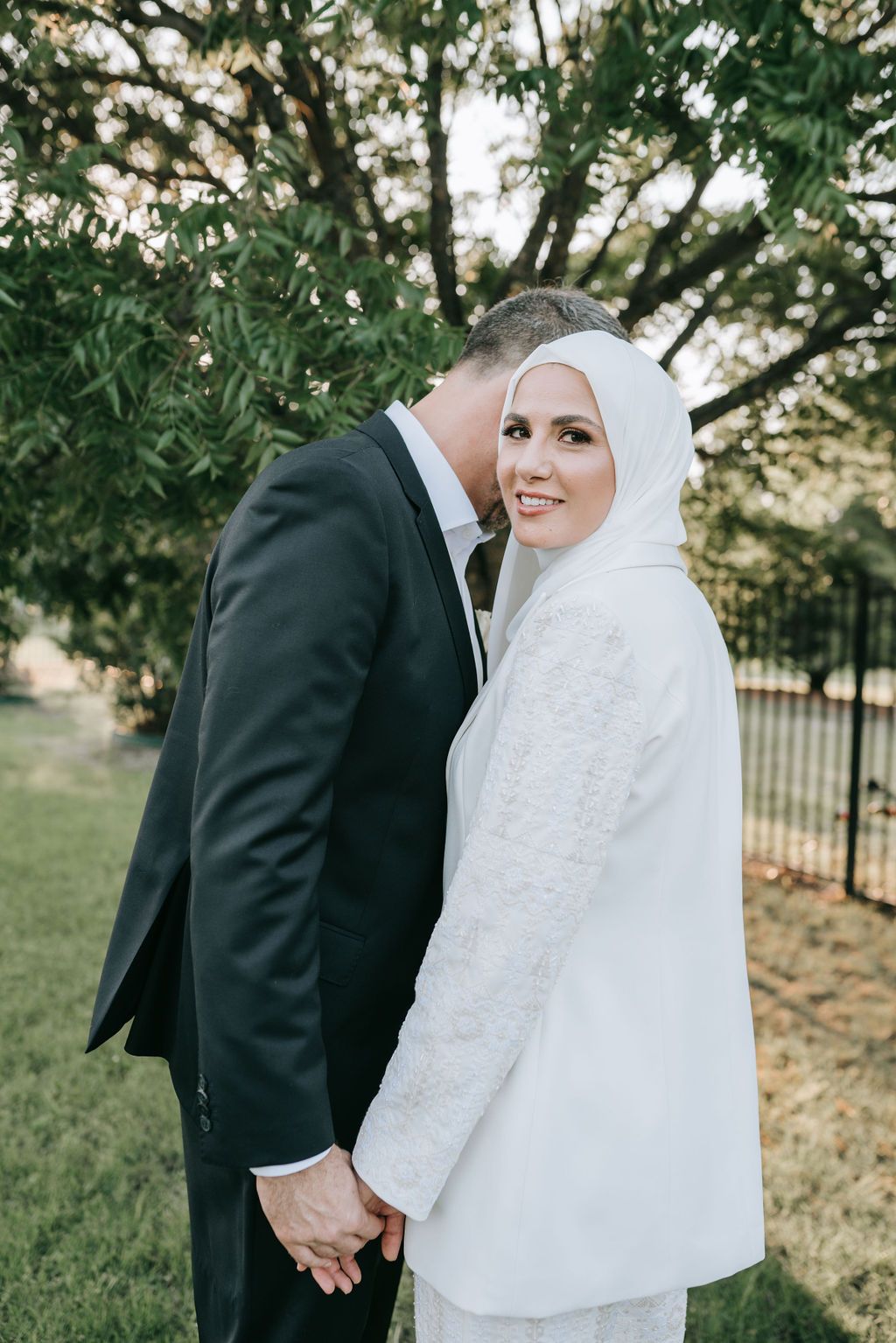 A wedding couple sharing an intimate moment in a garden setting, with the bride wearing an elegant white hijab.