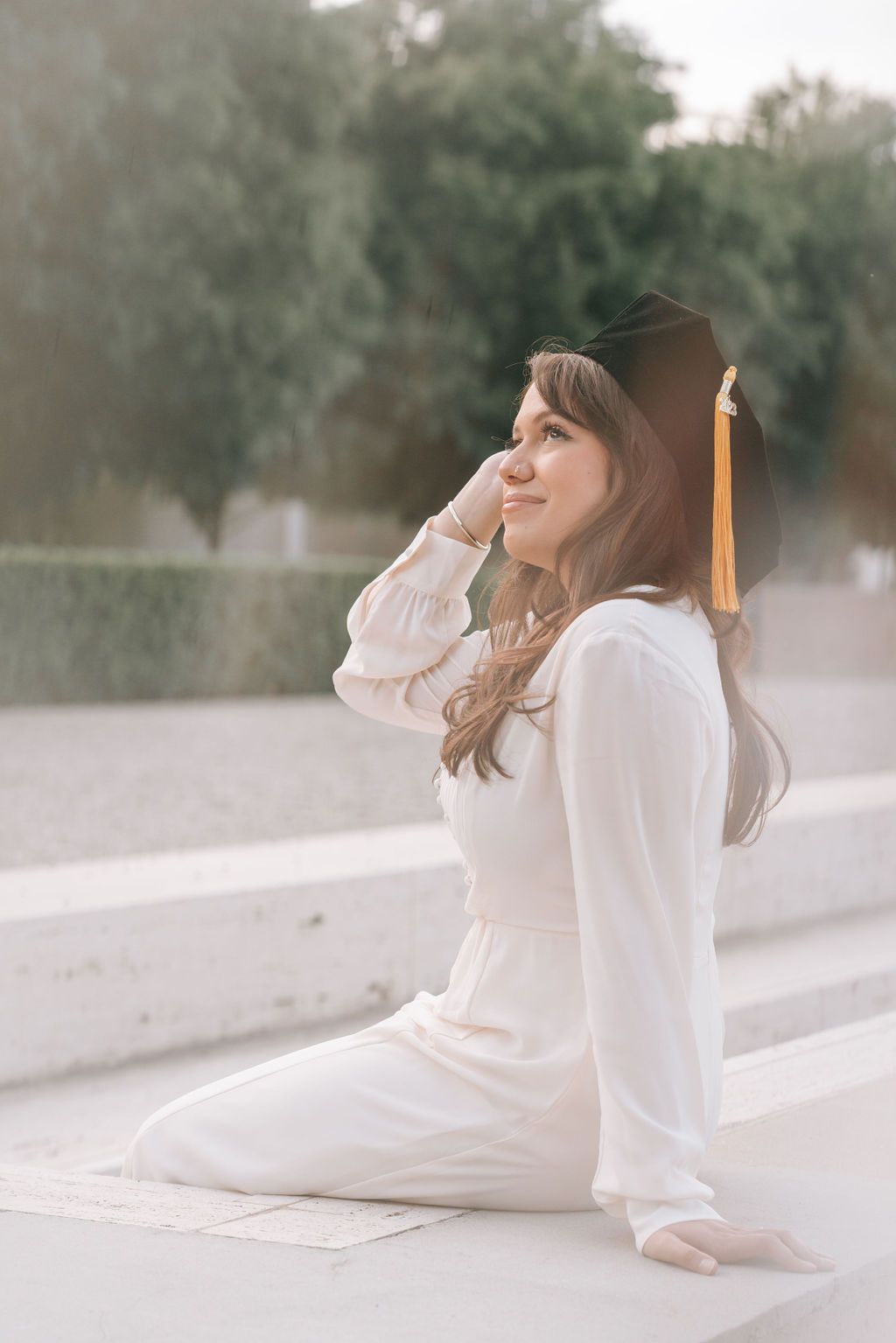 Graduate in white outfit sits reflectively during graduation photoshoot.