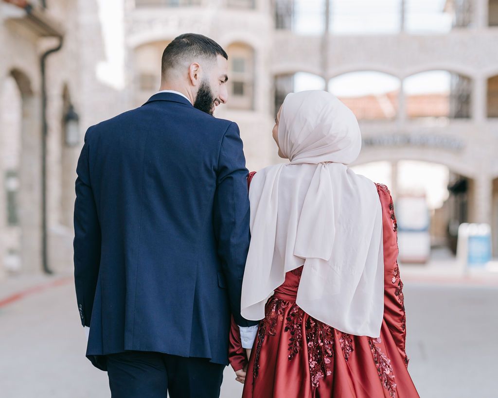 Couple in formal attire walking together, with one wearing a navy suit and the other in a red dress with white hijab.
