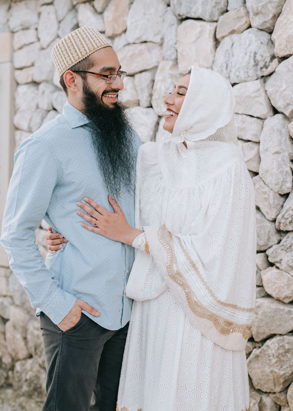 Couple in traditional Islamic dress share a tender moment against stone wall backdrop.
