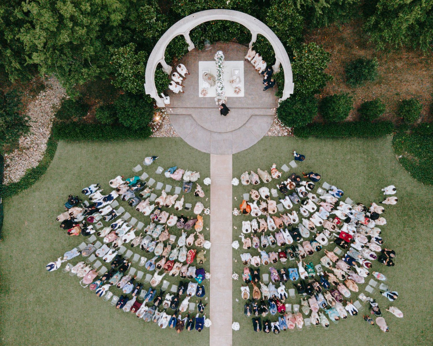 Aerial view of outdoor wedding ceremony with circular arch and rows of guests arranged in a fan pattern on green lawn.
