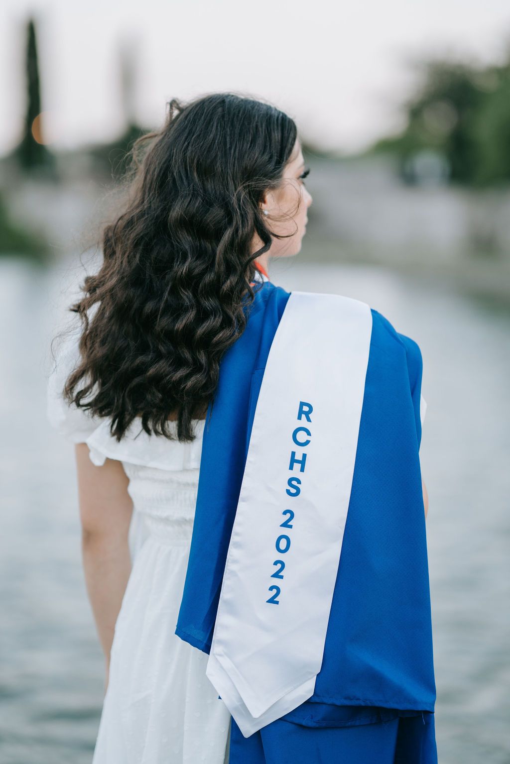 Graduate wearing white dress displays class of 2022 stole against blurred background.