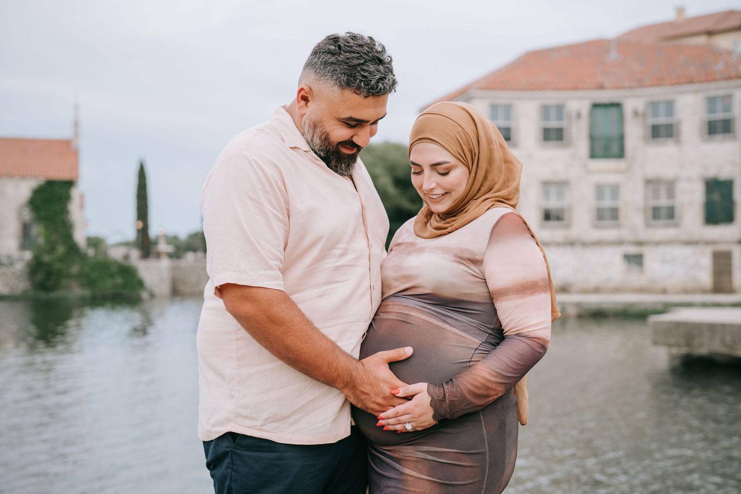 A couple tenderly holds a pregnant belly while standing by a waterfront with buildings in the background.