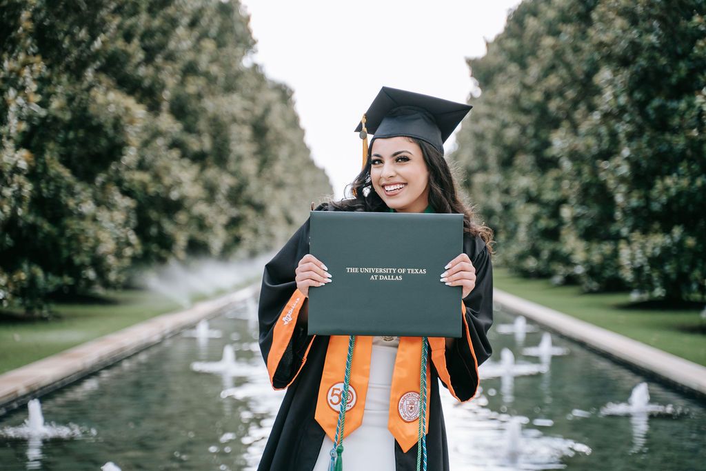 Graduate in cap and gown proudly displays diploma while standing near fountains in landscaped garden setting.